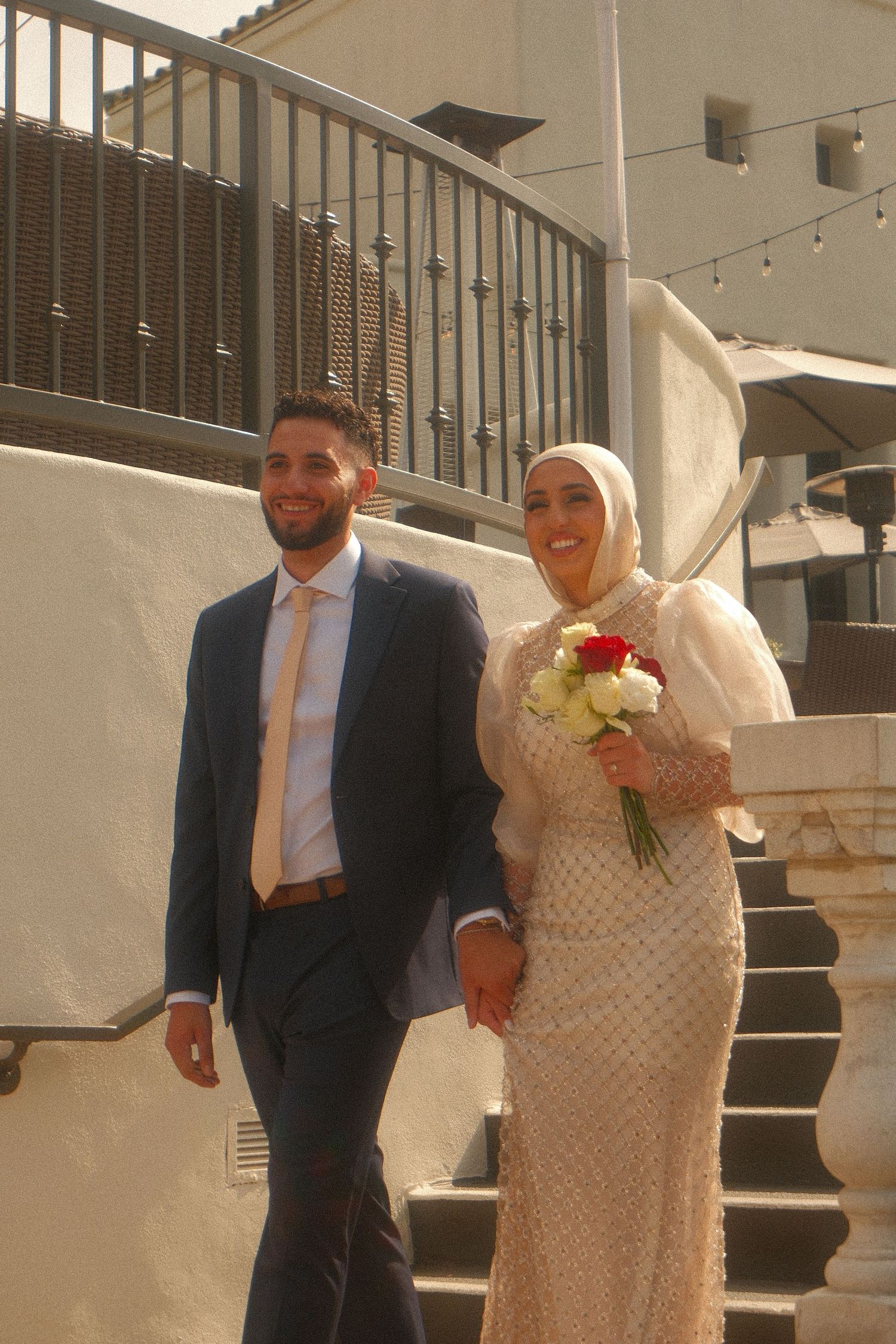 Smiling bride in hijab holding red and white bouquet walking with groom in navy suit on outdoor steps.