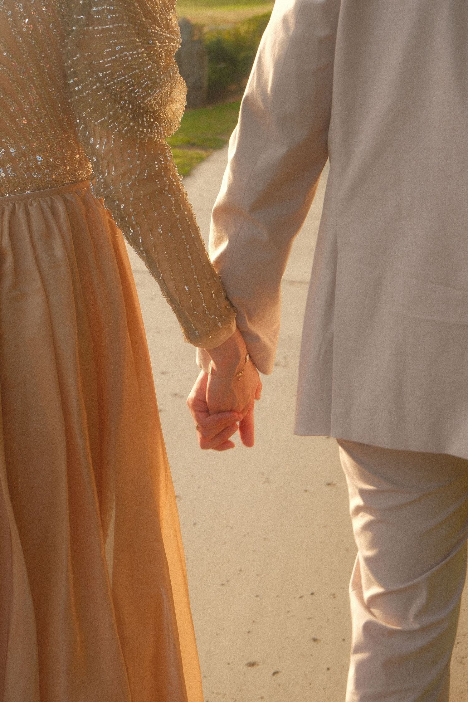 Couple holding hands at golden hour, woman in gold sequin gown and man in beige suit walking on path.