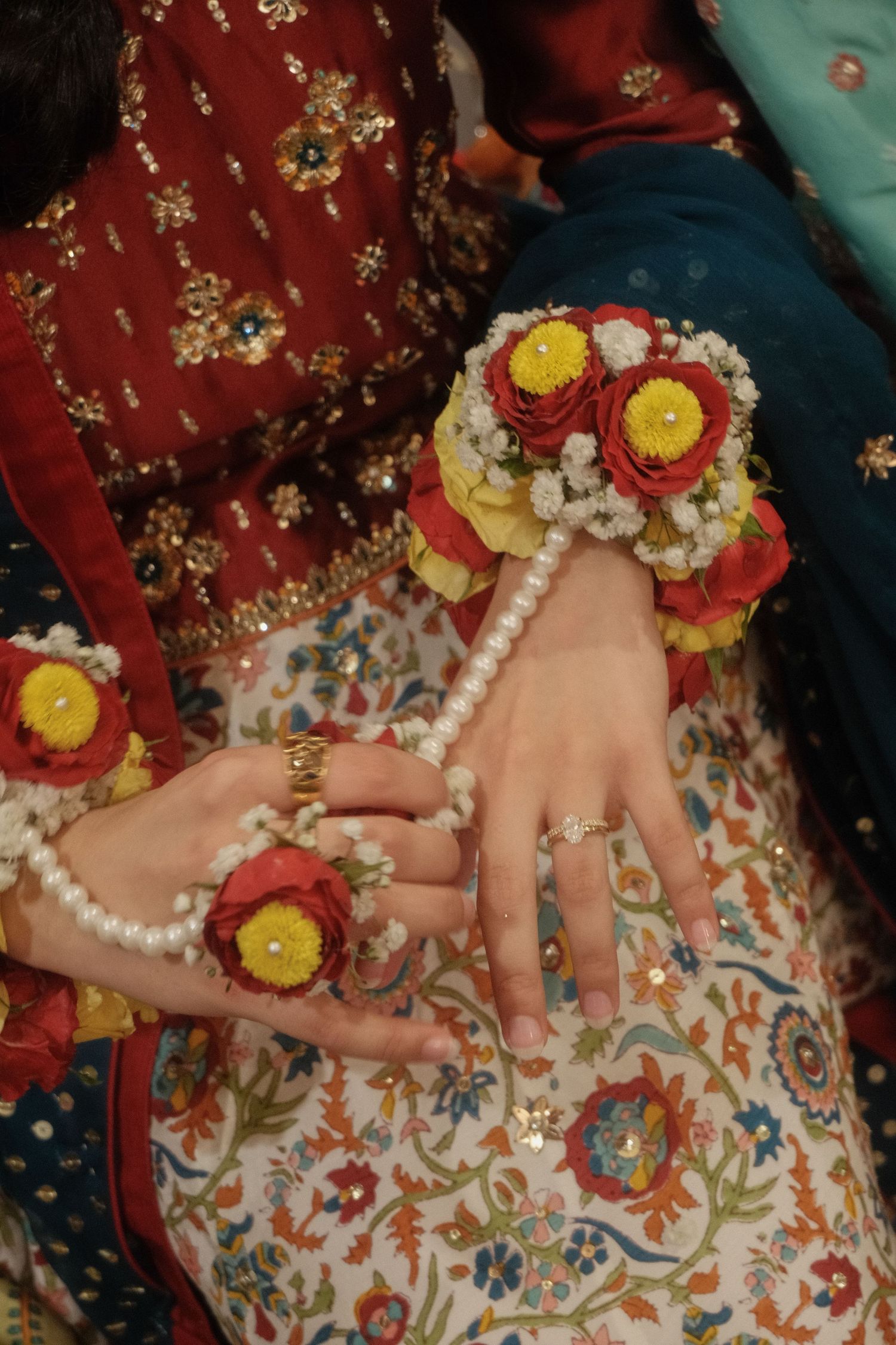 Bride wearing floral wrist corsage made of red and yellow flowers with pearl bracelet on white floral lehenga.