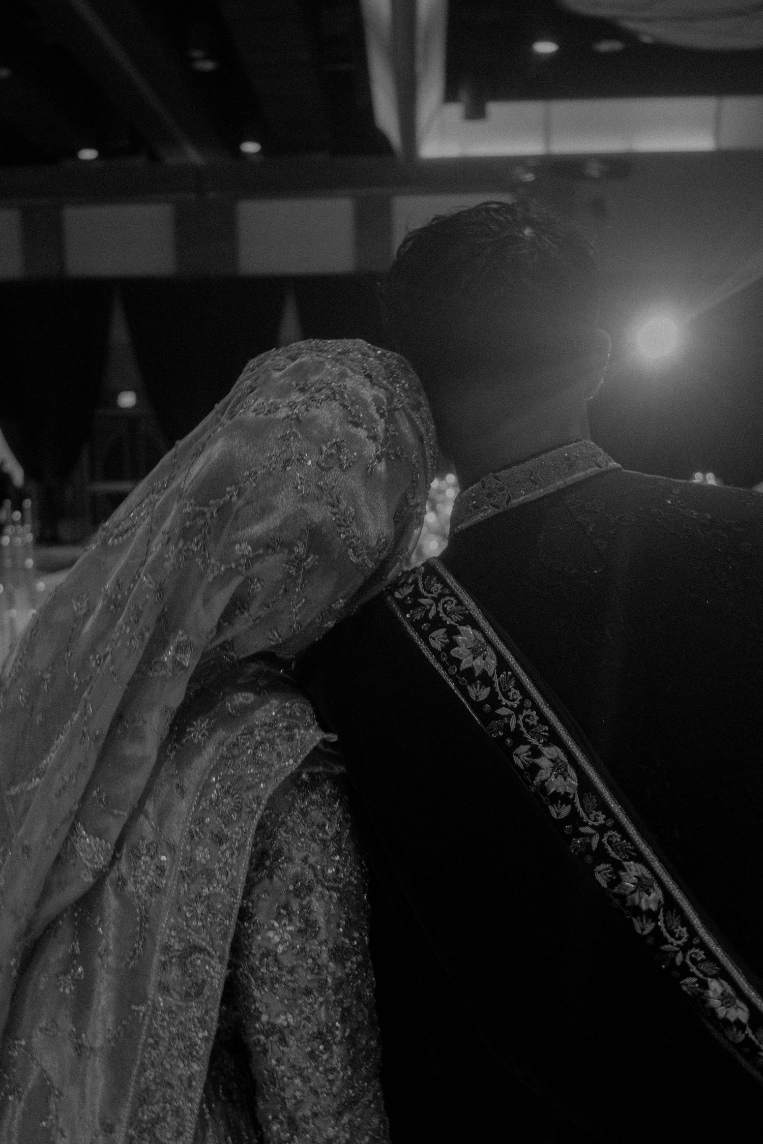 Black and white artistic photo of South Asian bride leaning on groom's shoulder in traditional wedding attire.