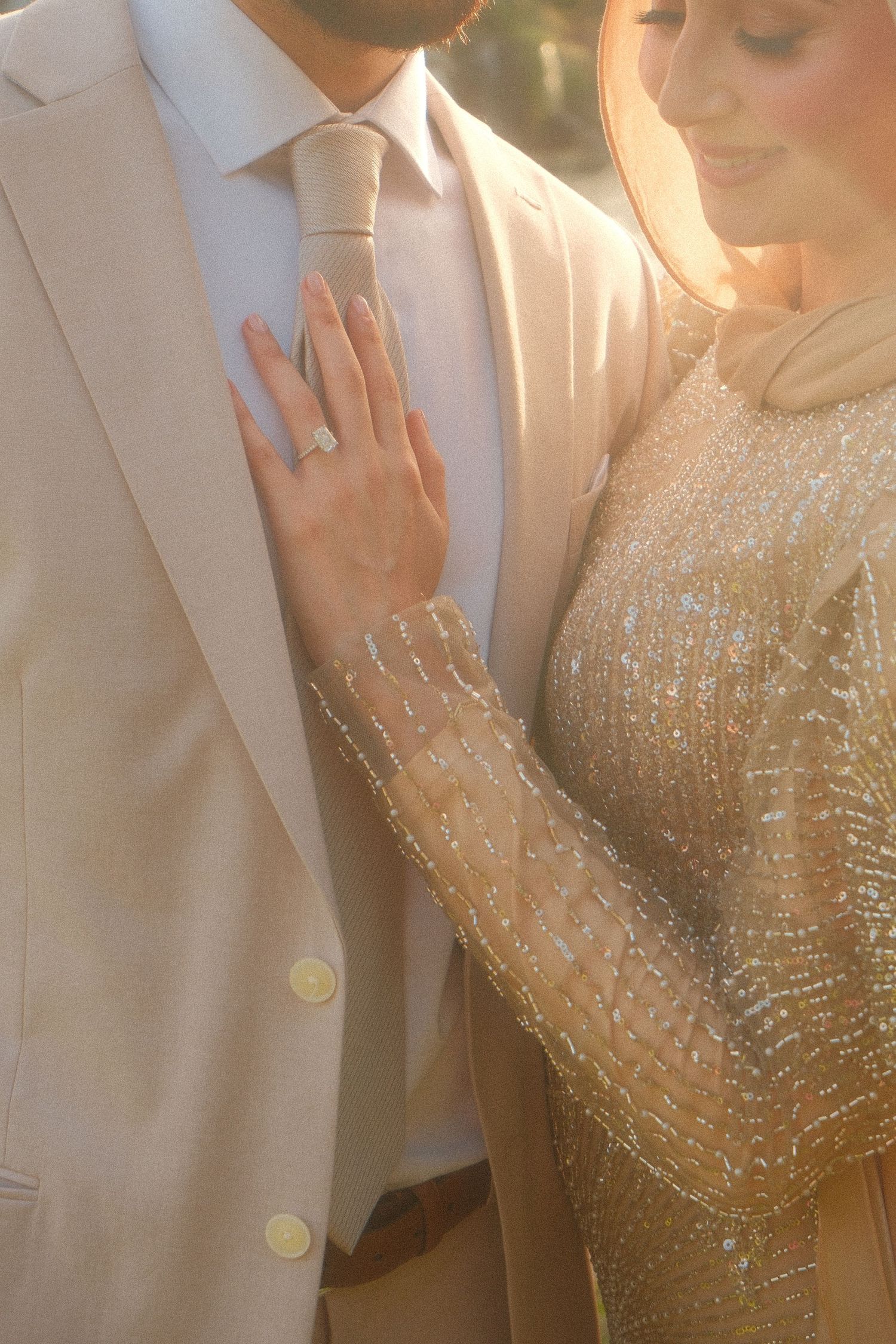 Close-up of bride in gold beaded gown showing diamond engagement ring while resting hand on groom's beige suit.
