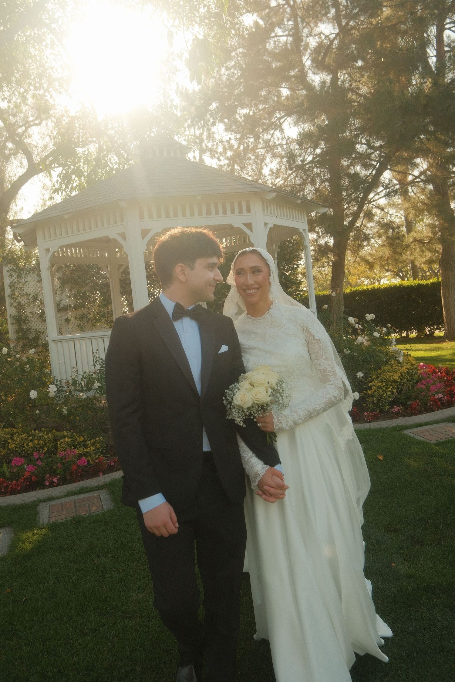 Bride and groom smiling together in a sunlit garden with a white gazebo and colorful flowers in the background.