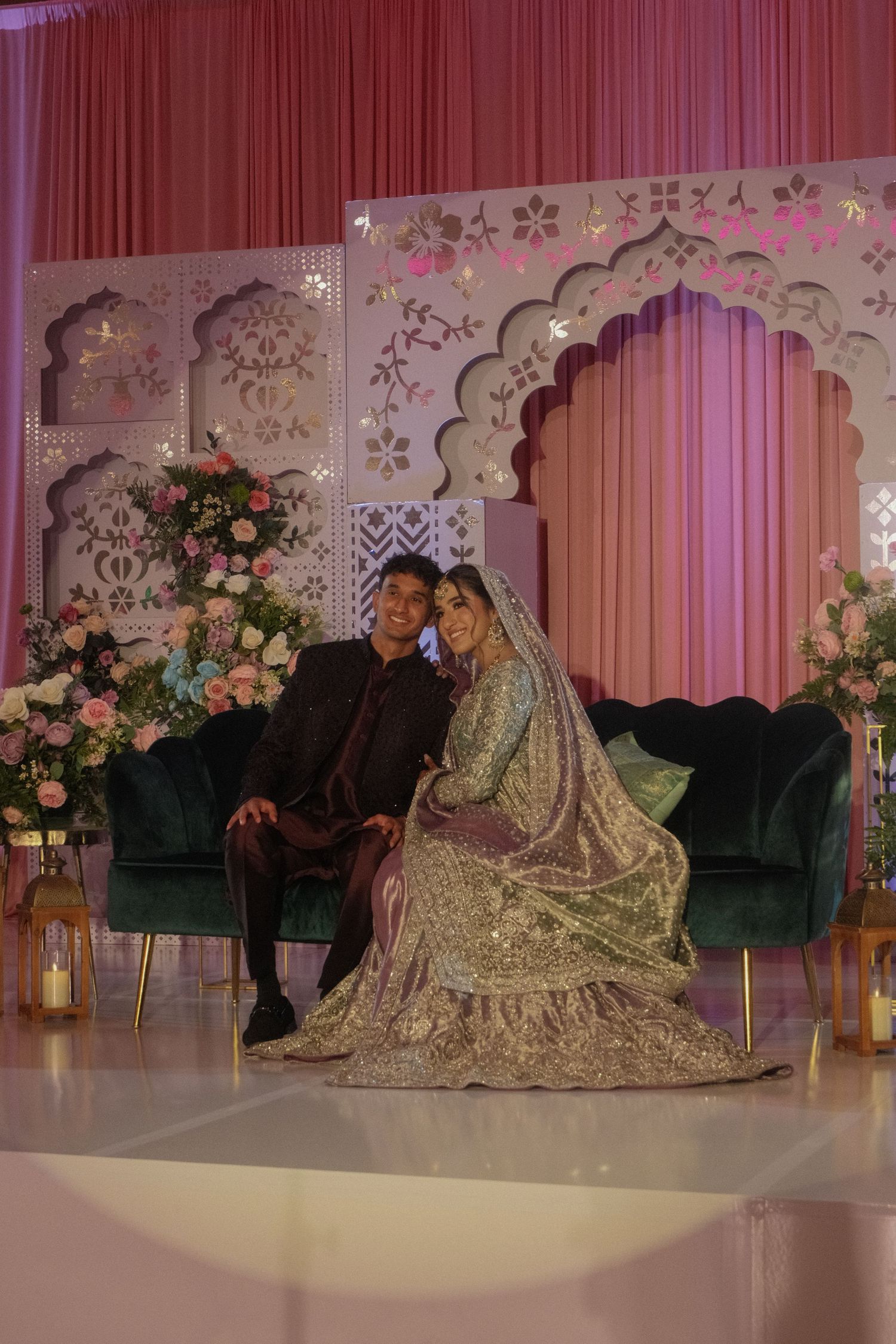 South Asian bride and groom seated together on green velvet sofa at decorated wedding stage.