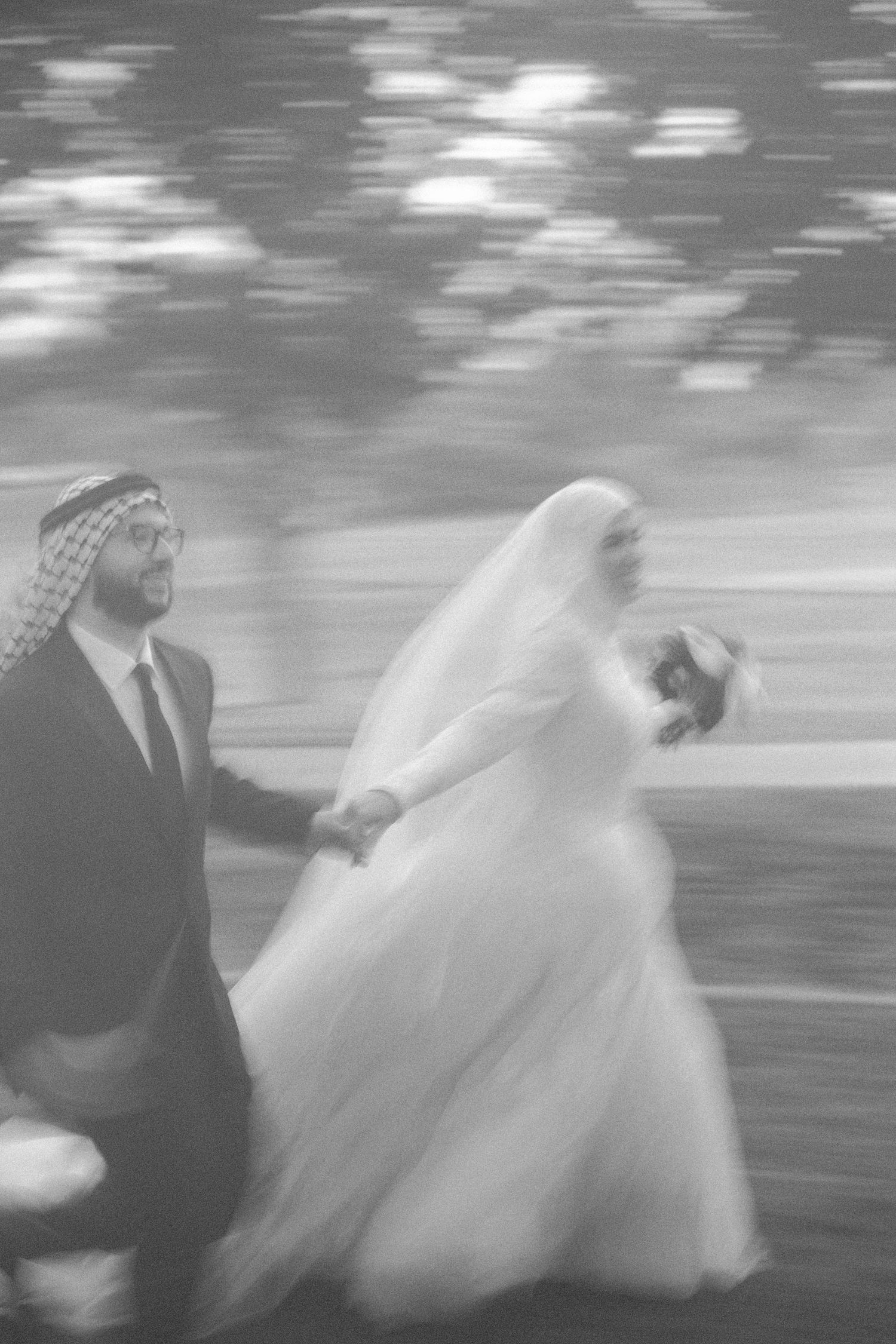 Black and white motion blur photo of Muslim wedding couple running hand in hand under trees.