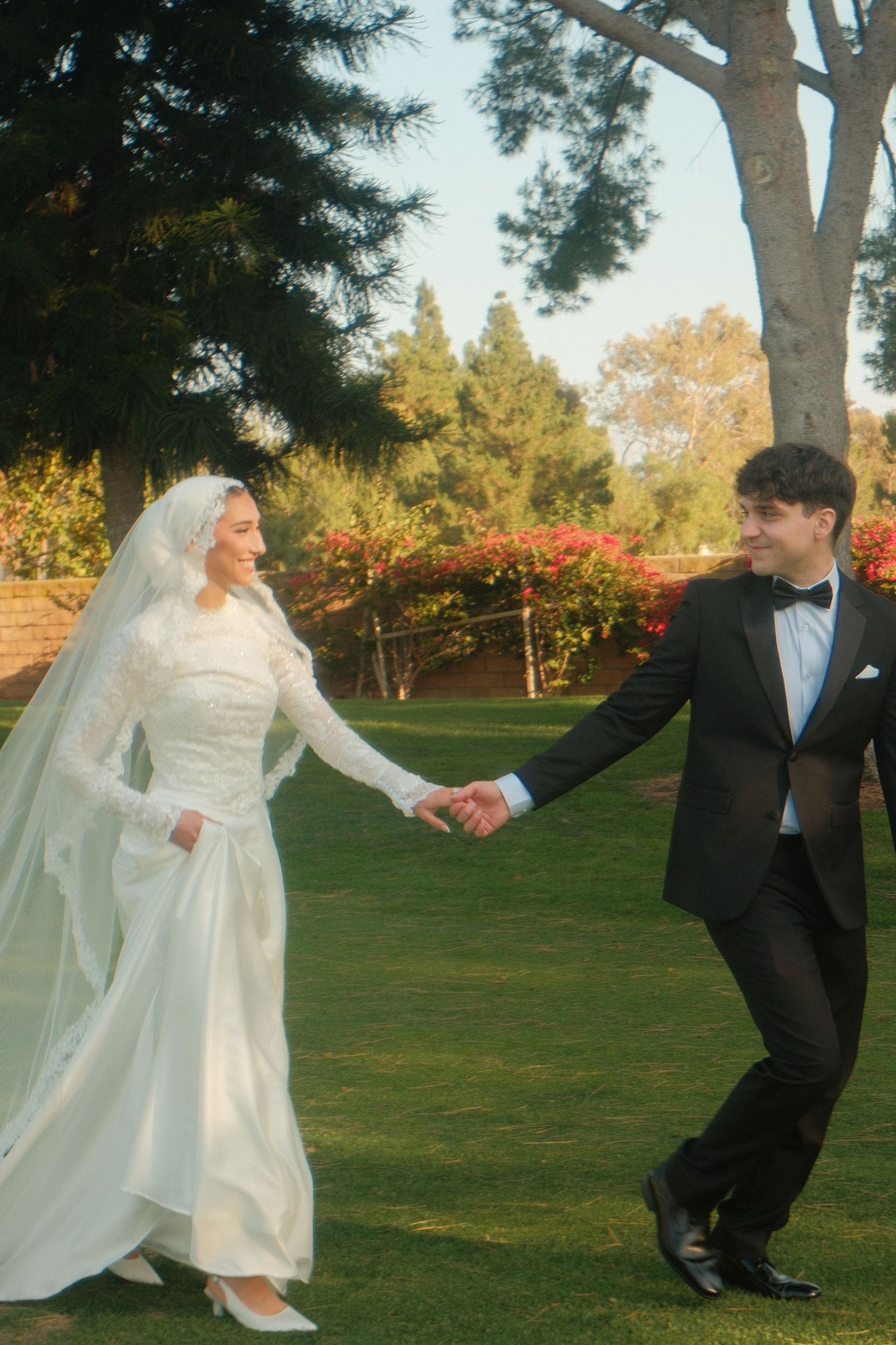 Newlyweds holding hands and walking in a lush garden, bride in white gown with veil, groom in tuxedo.