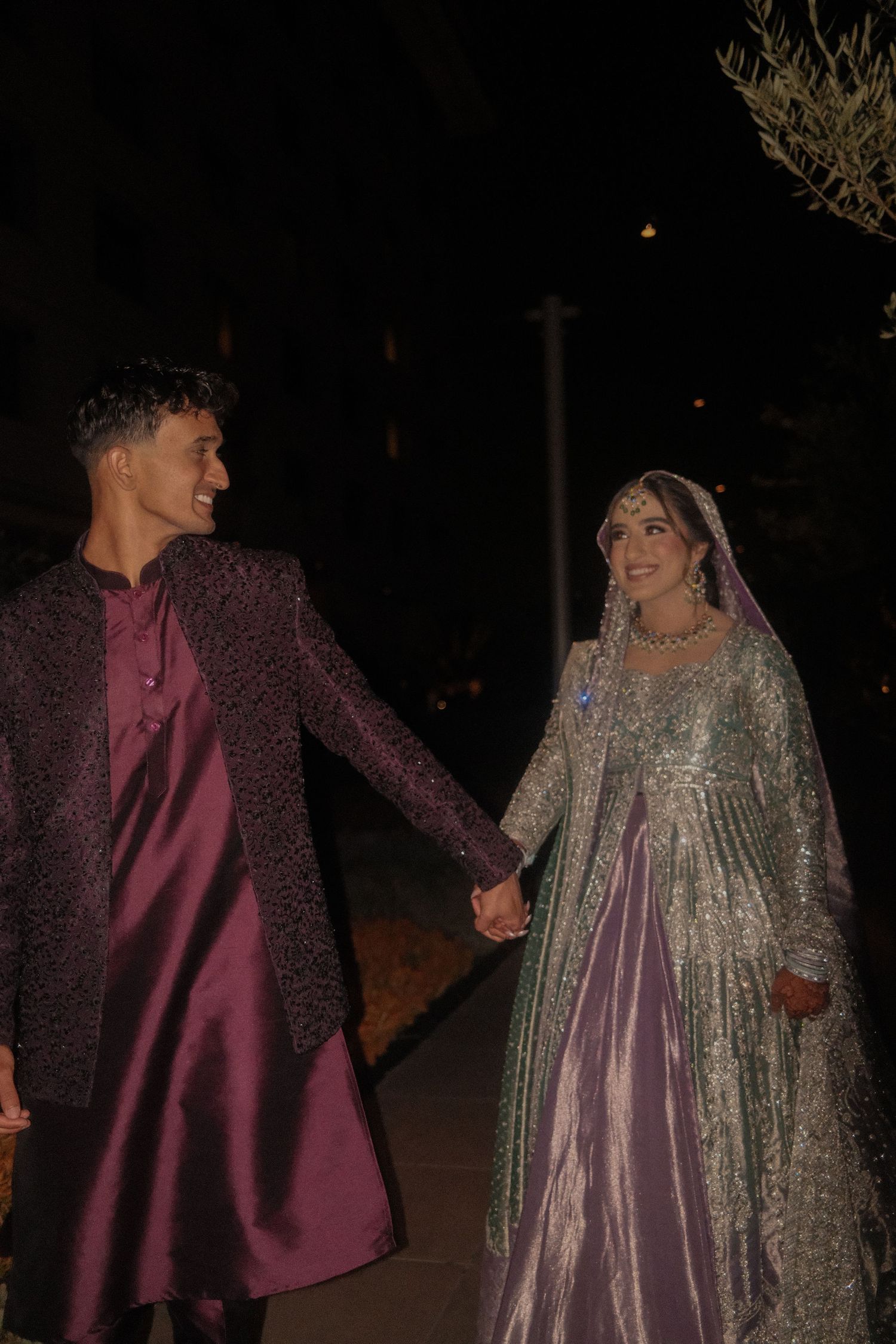 South Asian wedding couple holding hands and smiling at night, groom in maroon sherwani bride in silver lehenga.