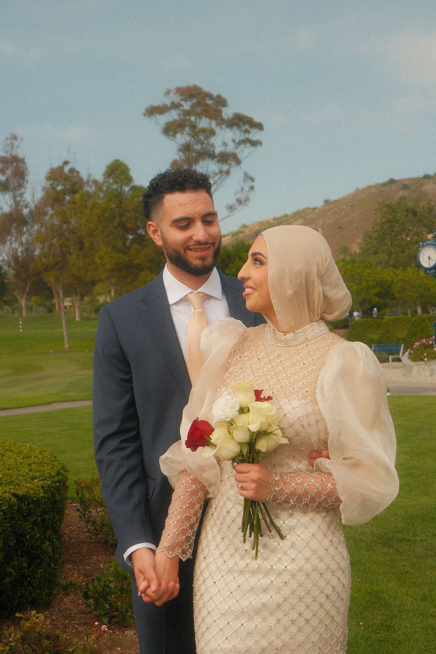 Bride in beige hijab and beaded gown holding bouquet gazing at groom in navy suit on green lawn.