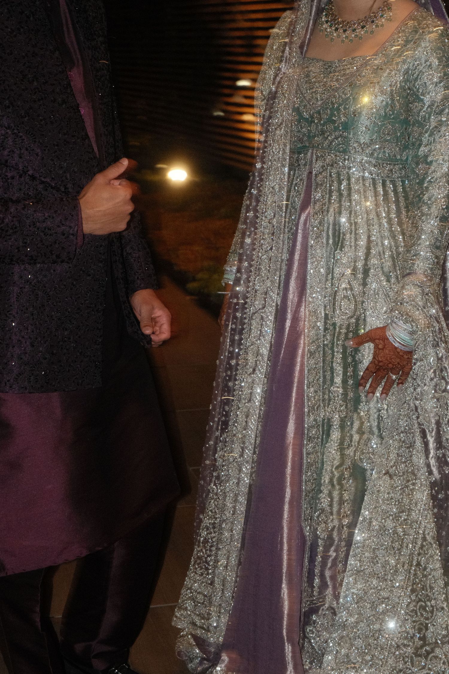 South Asian couple in traditional wedding attire, groom in dark sherwani, bride in silver embroidered lehenga with dupatta.