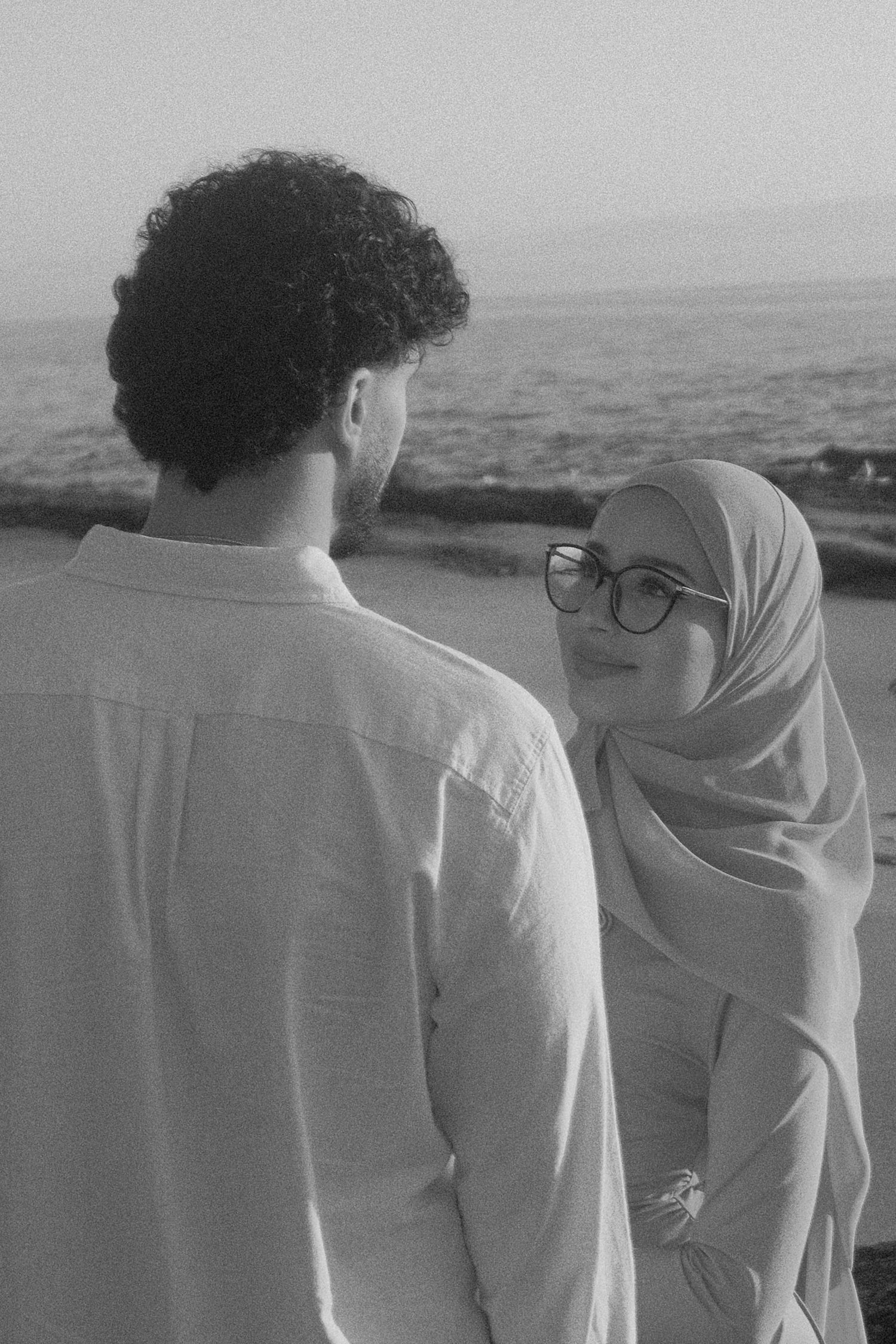 Monochrome seaside portrait of a couple, hijab-wearing woman with glasses looking at her partner.