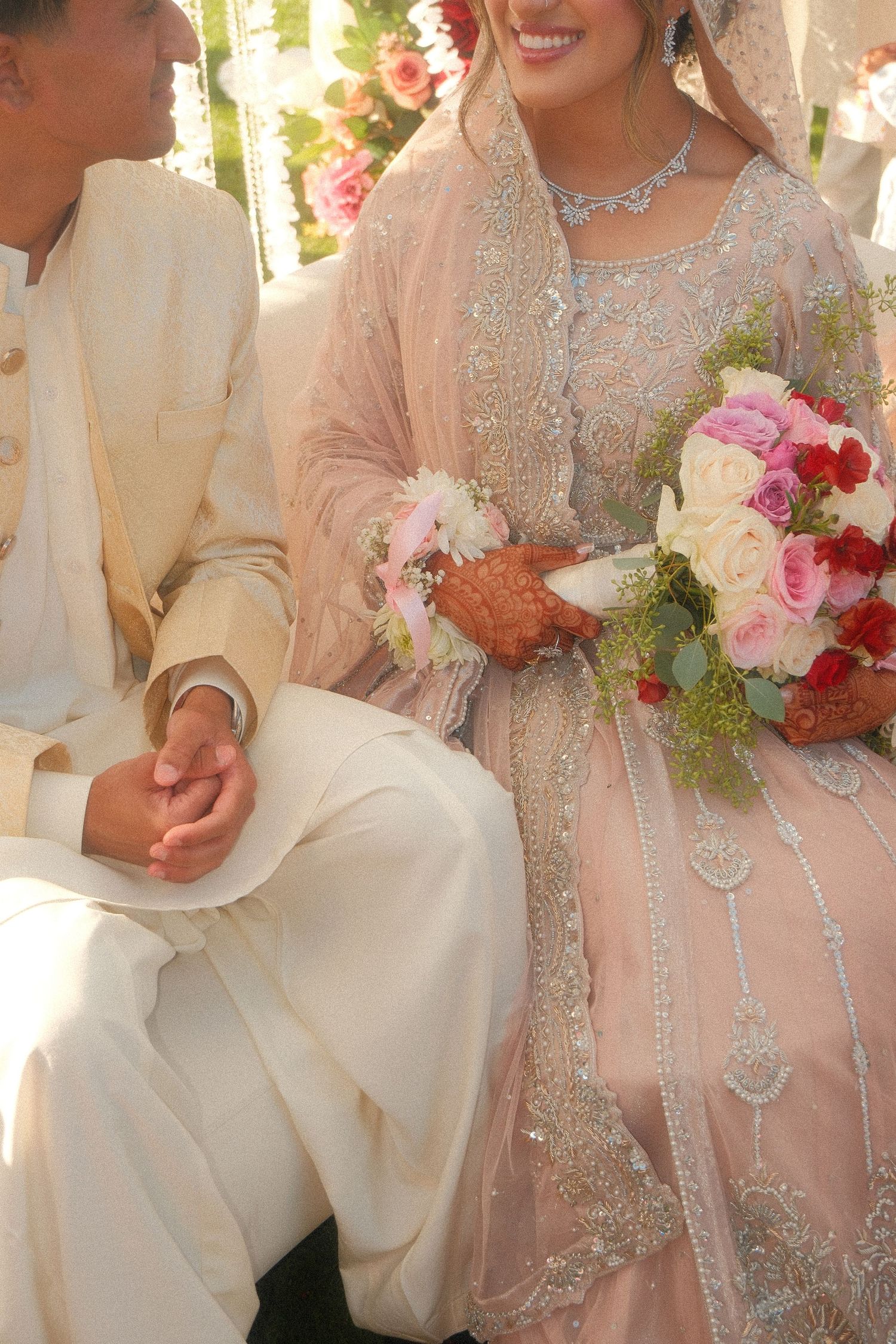 Muslim bride in embroidered blush pink outfit holding a colorful bouquet while sitting with groom.