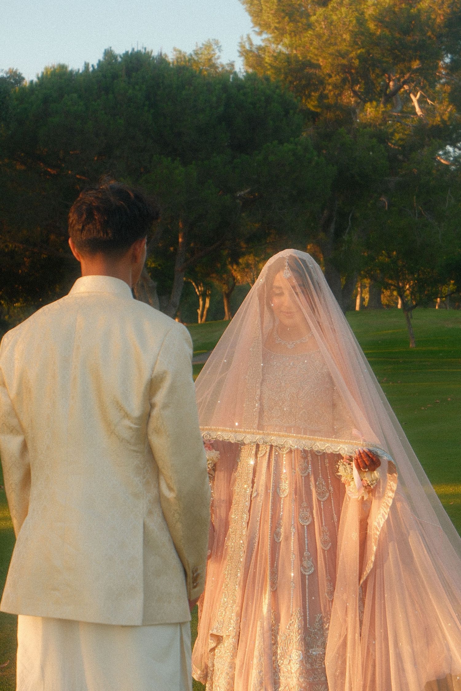 South Asian wedding couple from behind in garden at sunset, bride in embroidered lehenga with veil, groom in cream suit.