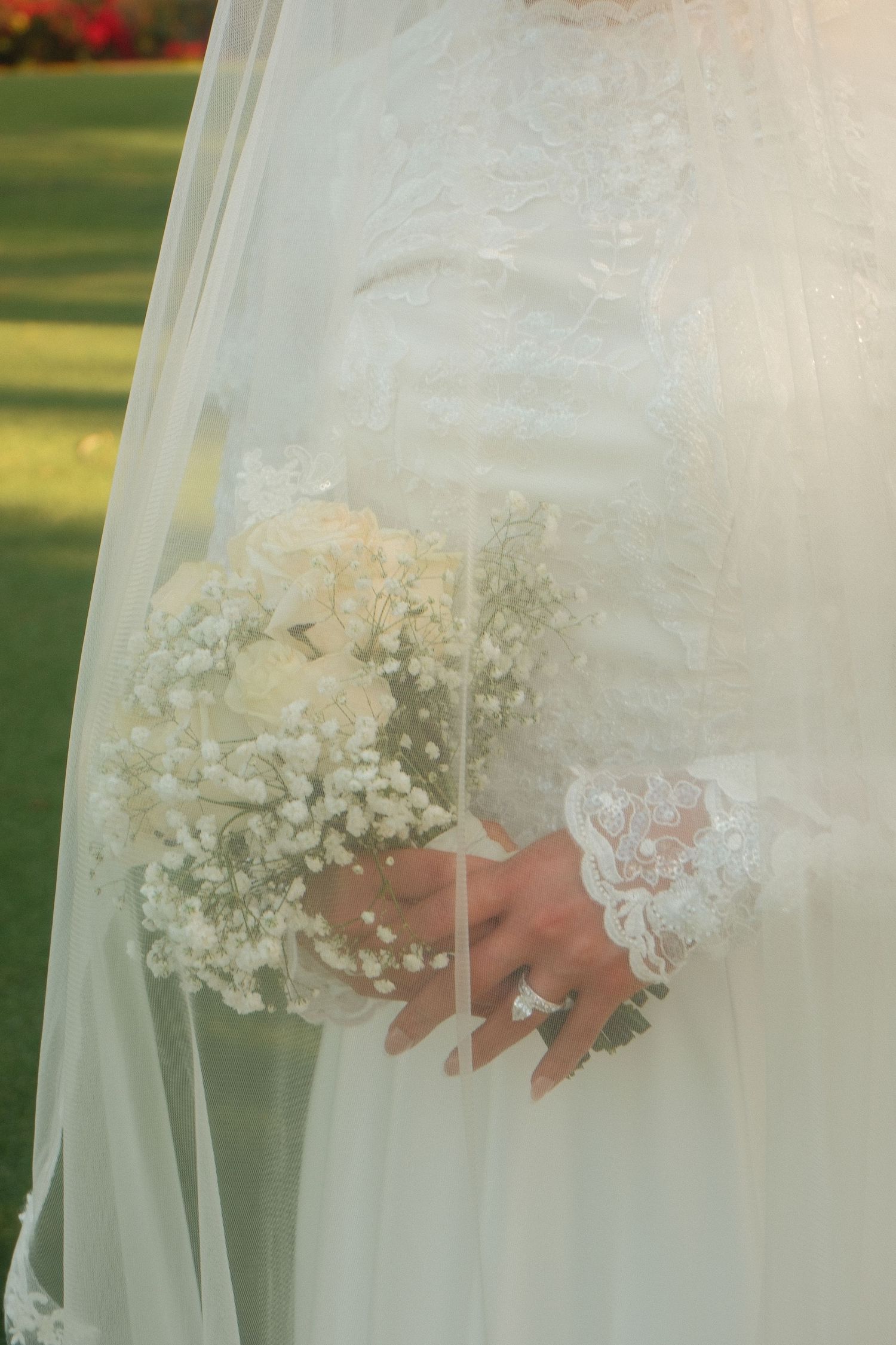 Bride in white lace wedding dress holding white rose and baby's breath bouquet with diamond ring outdoors.