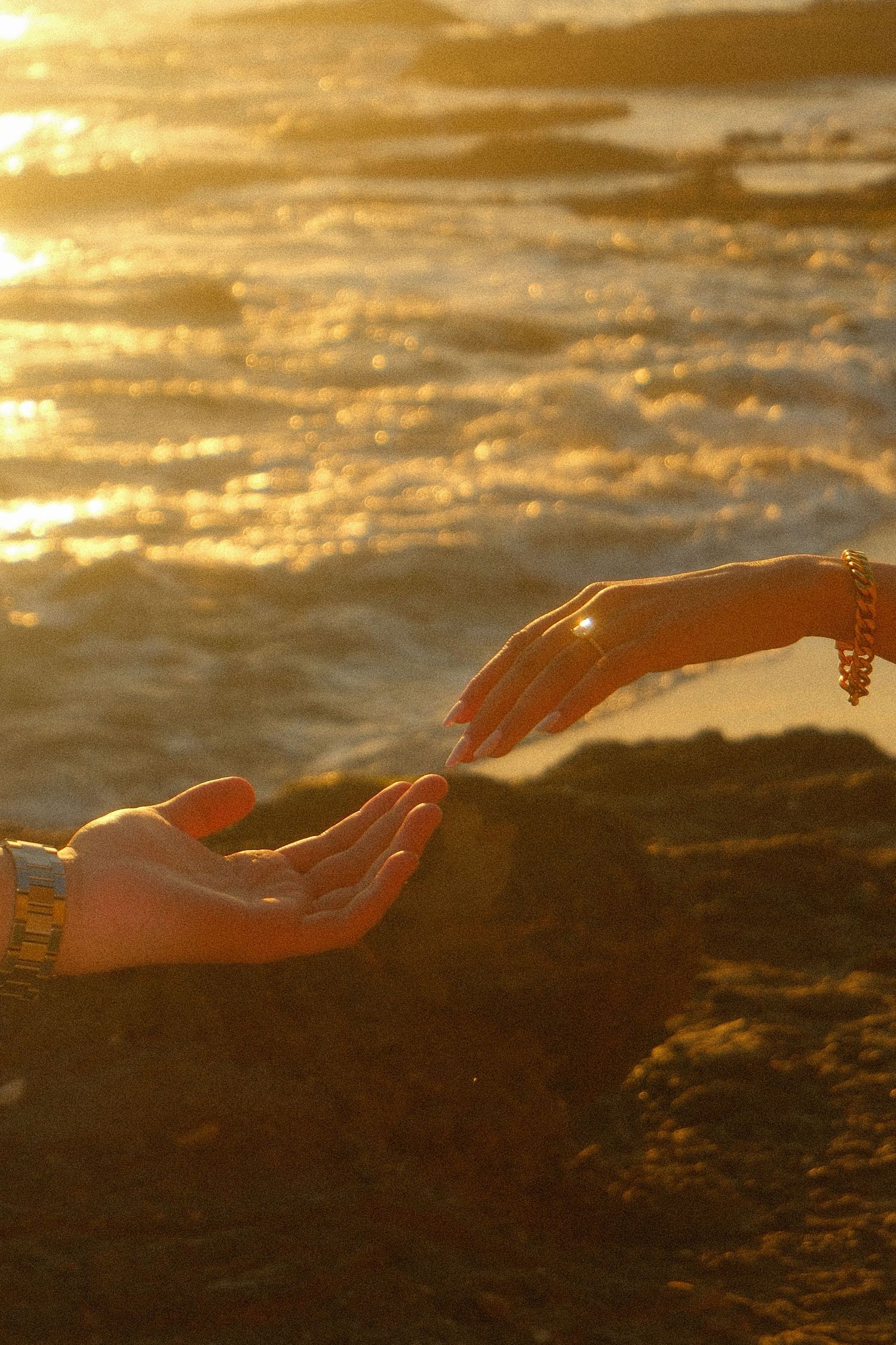 Two hands reaching toward each other at golden sunset on rocky ocean shoreline.