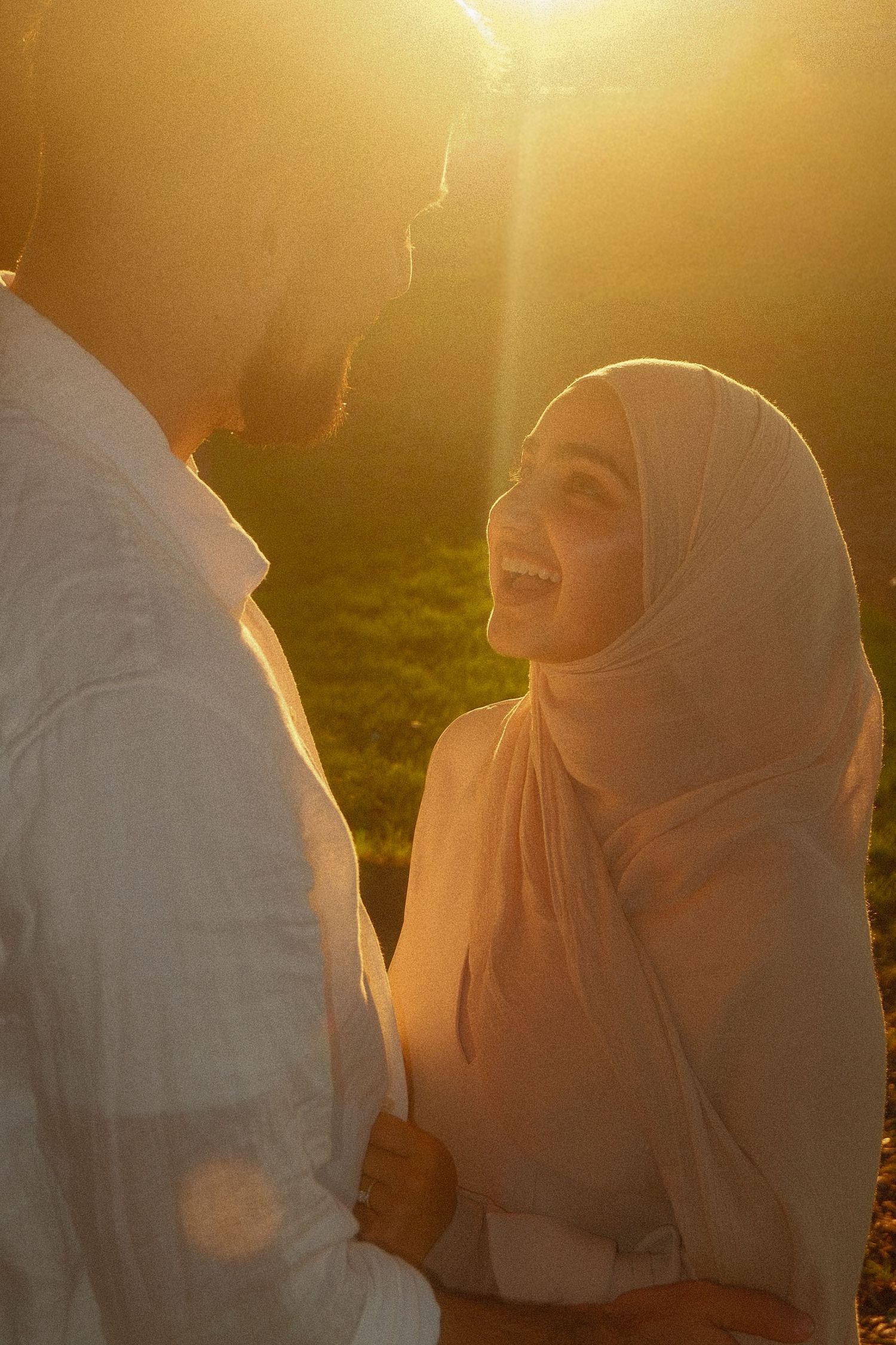 A joyful bride in a hijab laughs with her groom during a golden sunset outdoor photoshoot.
