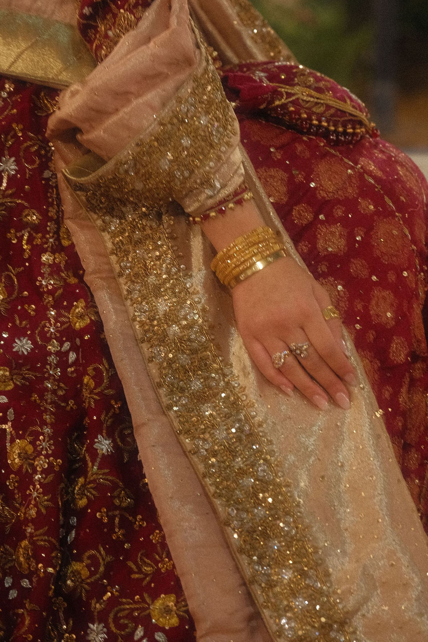 Bride in red gold Pakistani bridal attire showing diamond engagement ring and gold bangles on dupatta.