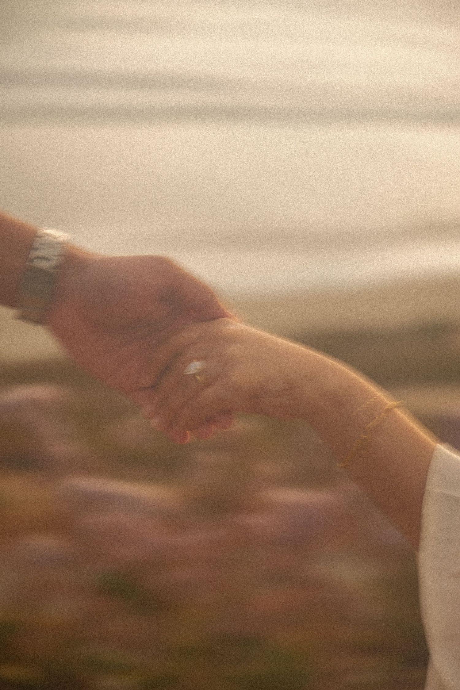 Couple holding hands at sunset near water, woman wearing diamond engagement ring and delicate bracelet.