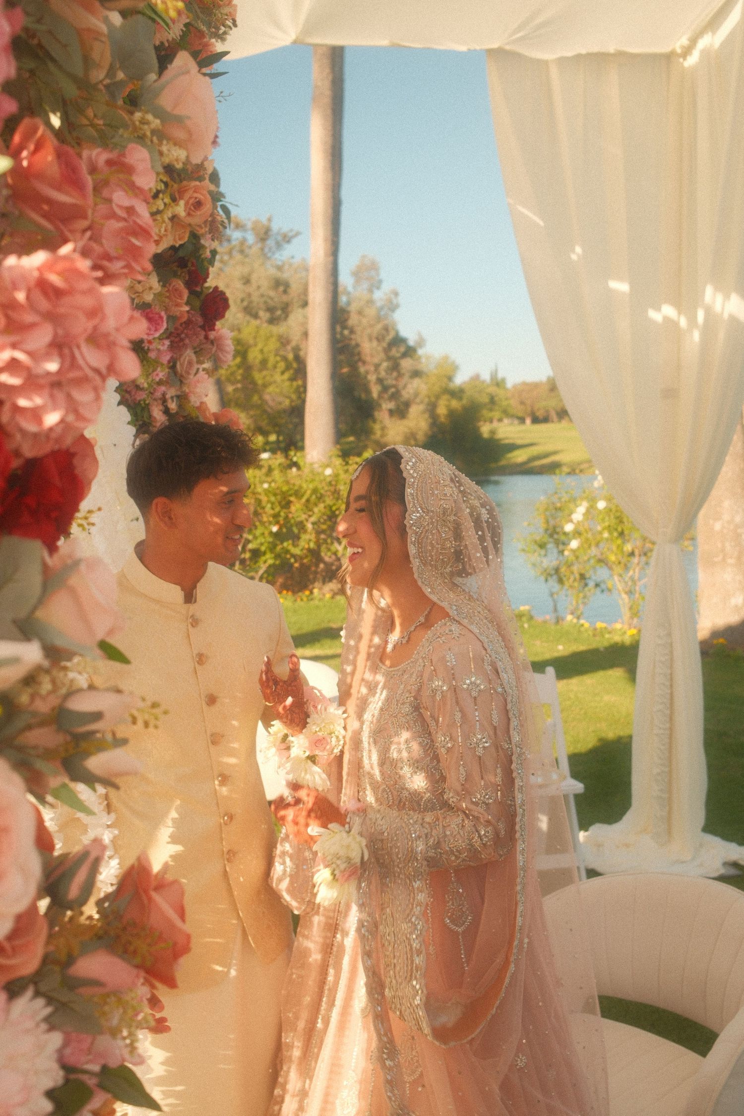 South Asian bride and groom in ivory wedding attire stand under floral arch with pink blooms outdoors.