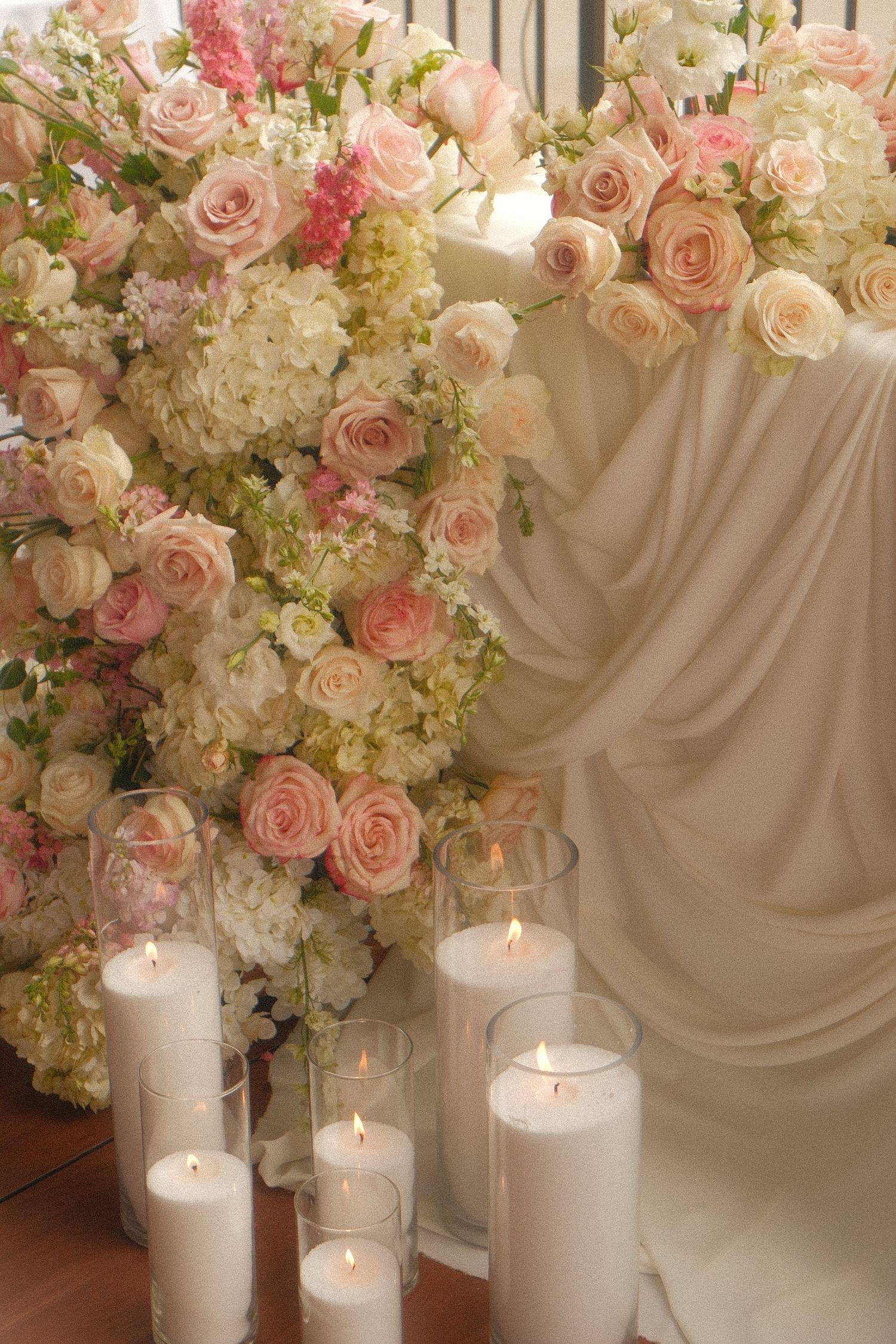 Elegant wedding decor with pink roses, white hydrangeas, and lit pillar candles in glass holders against draped white fabric.