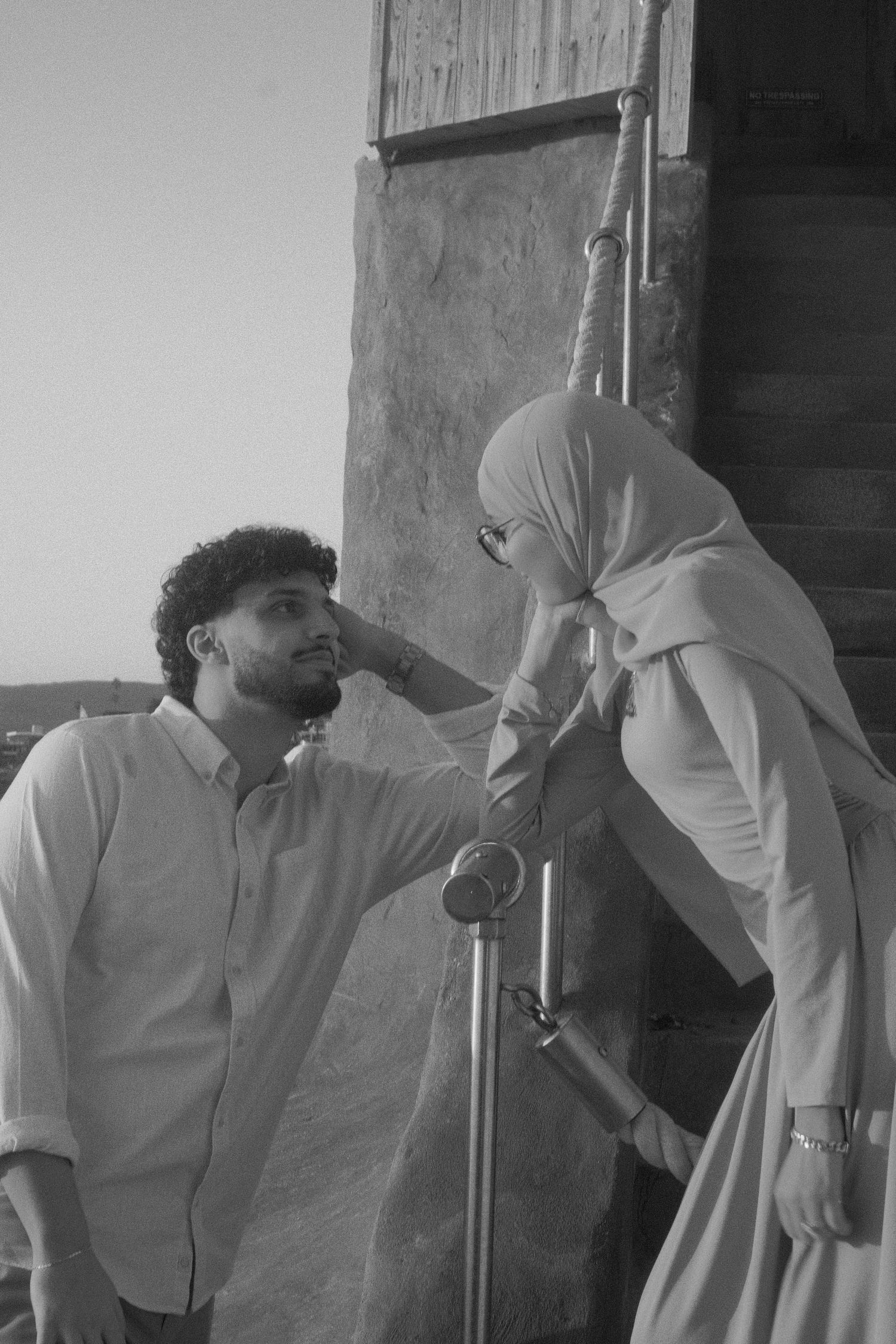 Black and white photo of a woman in hijab touching a bearded man's face outdoors near a stone wall.