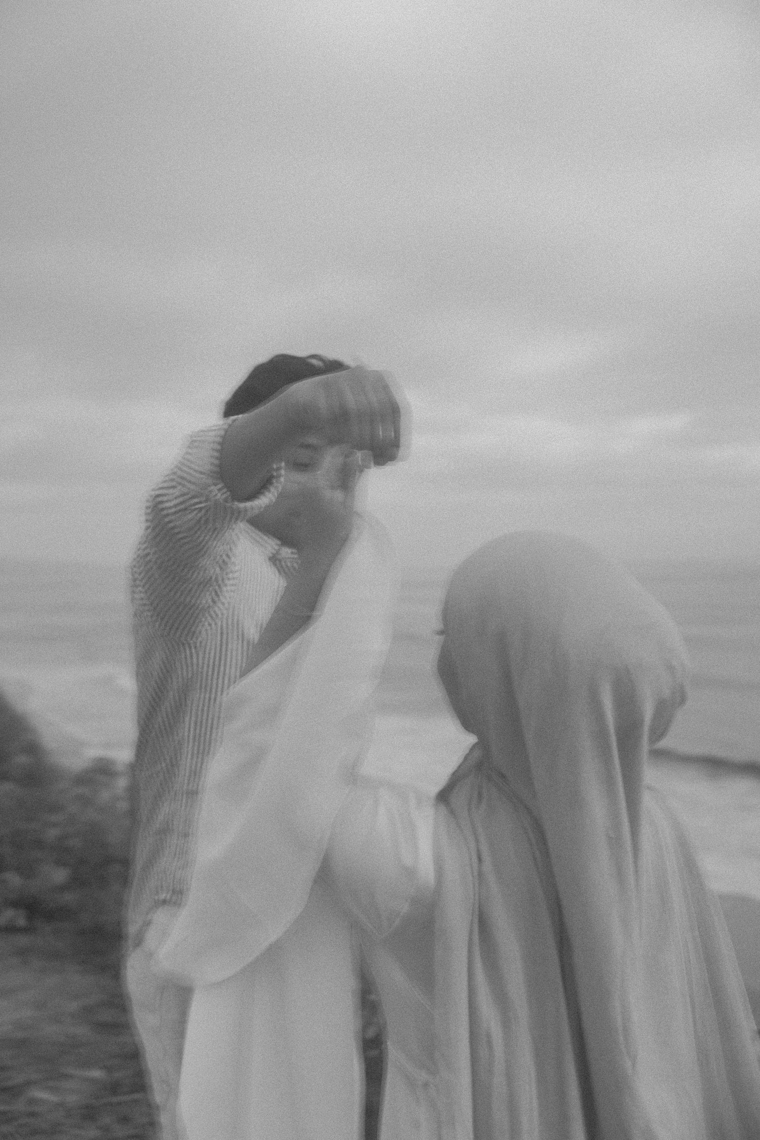 Black and white photo of a groom lifting a veil over his bride wearing a hijab at a seaside ceremony.
