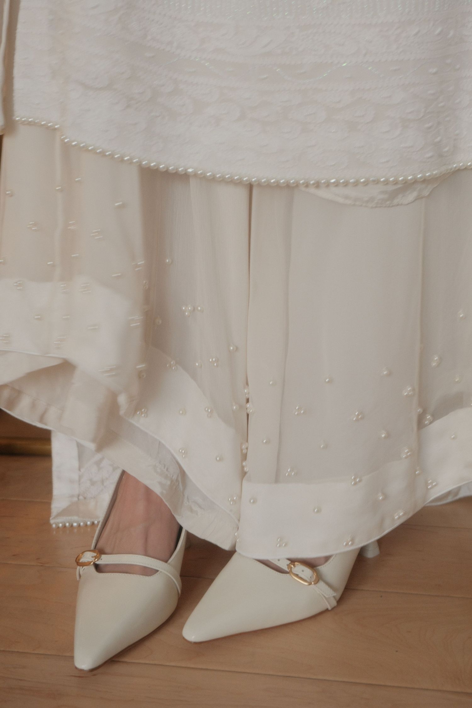 Close-up of bride wearing white pearl-embellished lehenga with pointed cream kitten heels on wooden floor.