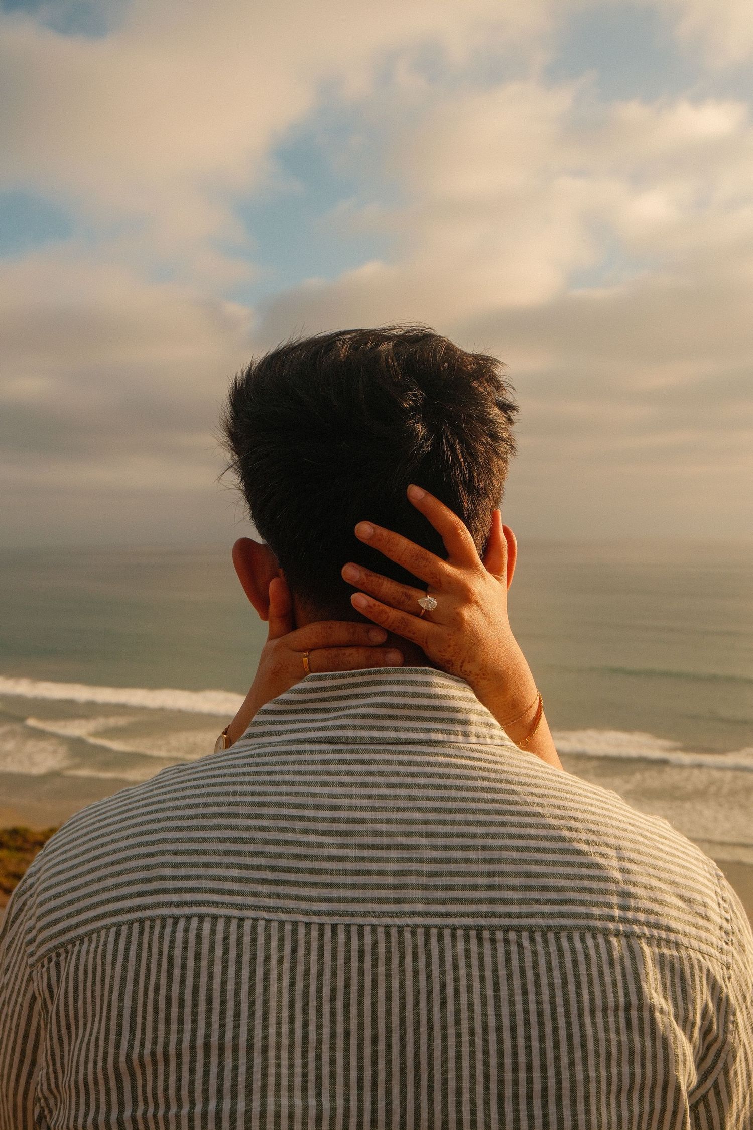 Woman placing hand on mans neck at beach sunset, engagement ring visible against striped linen shirt.