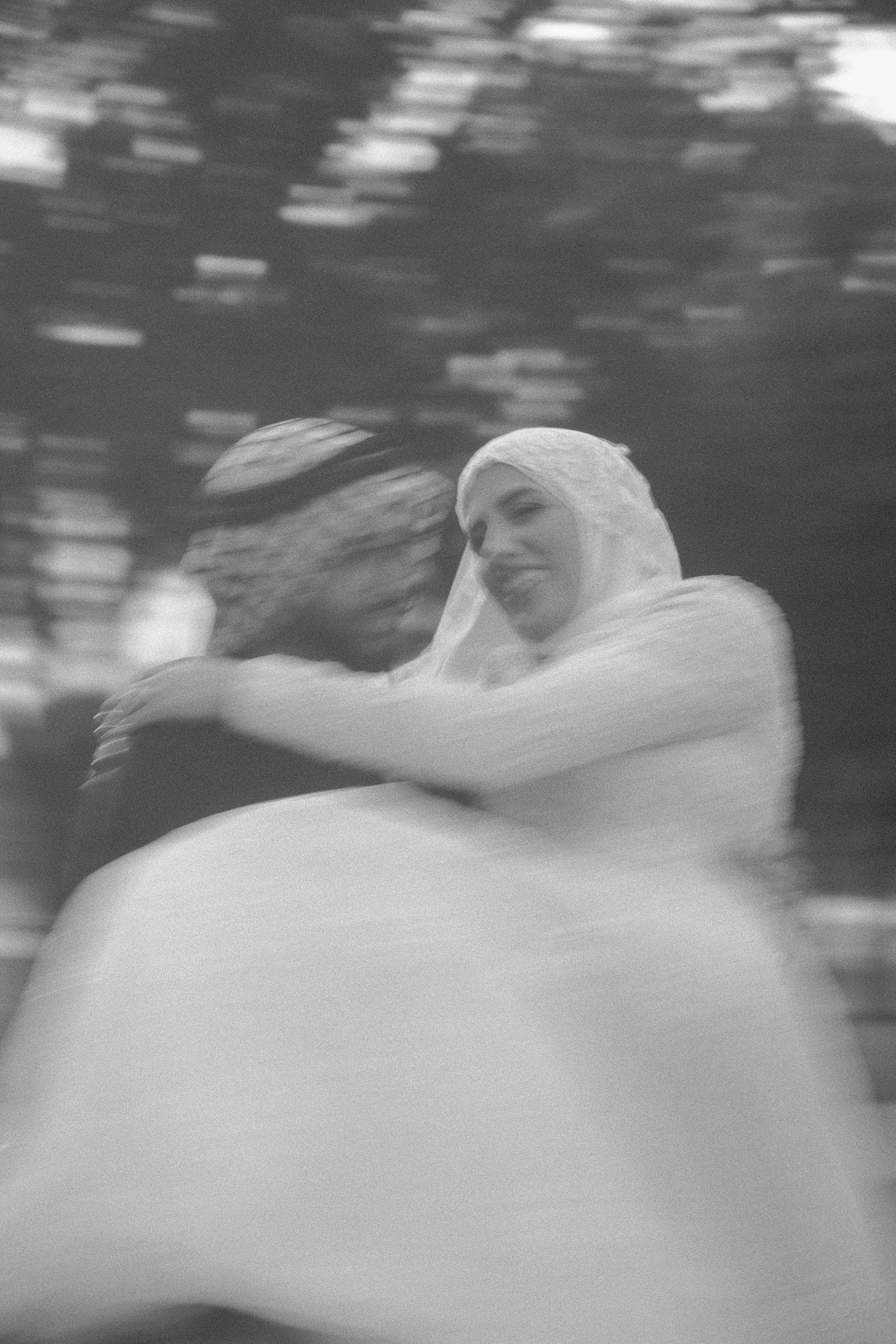 Black and white motion-blurred photo of Muslim wedding couple embracing, bride in white hijab.