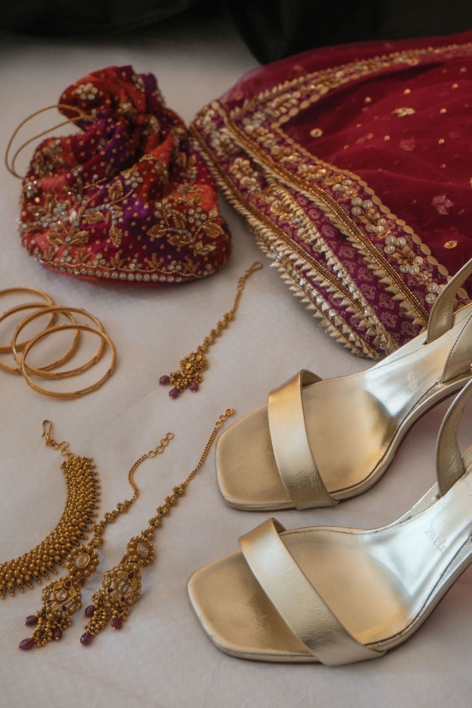 Gold bridal accessories including heels, bangles, necklace, earrings, and embroidered red potli bag on white surface.
