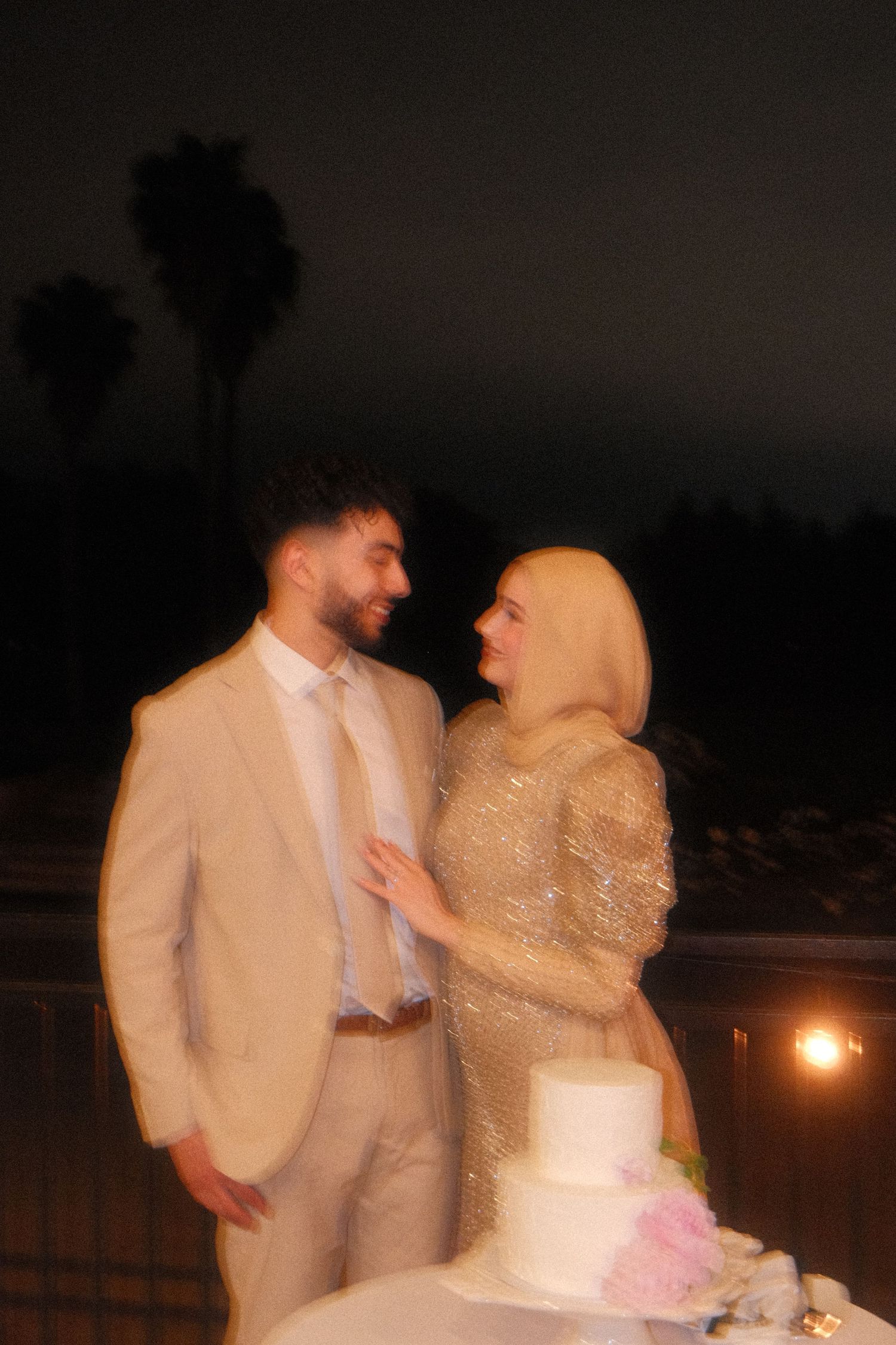 Couple sharing romantic moment by wedding cake at night, man in tan suit and woman in gold sequin dress.
