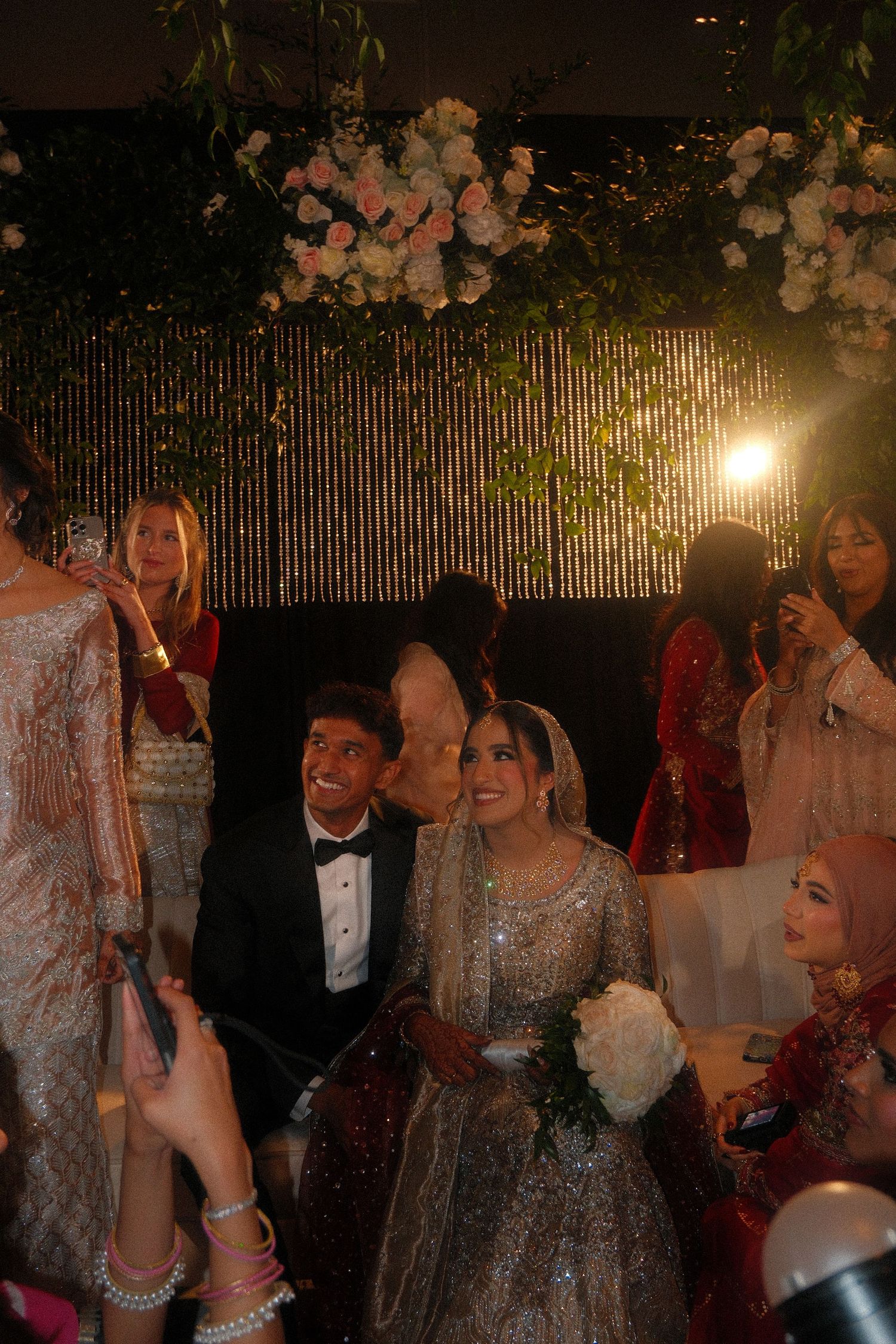 Pakistani wedding reception with smiling bride and groom seated on stage surrounded by guests and floral decorations.
