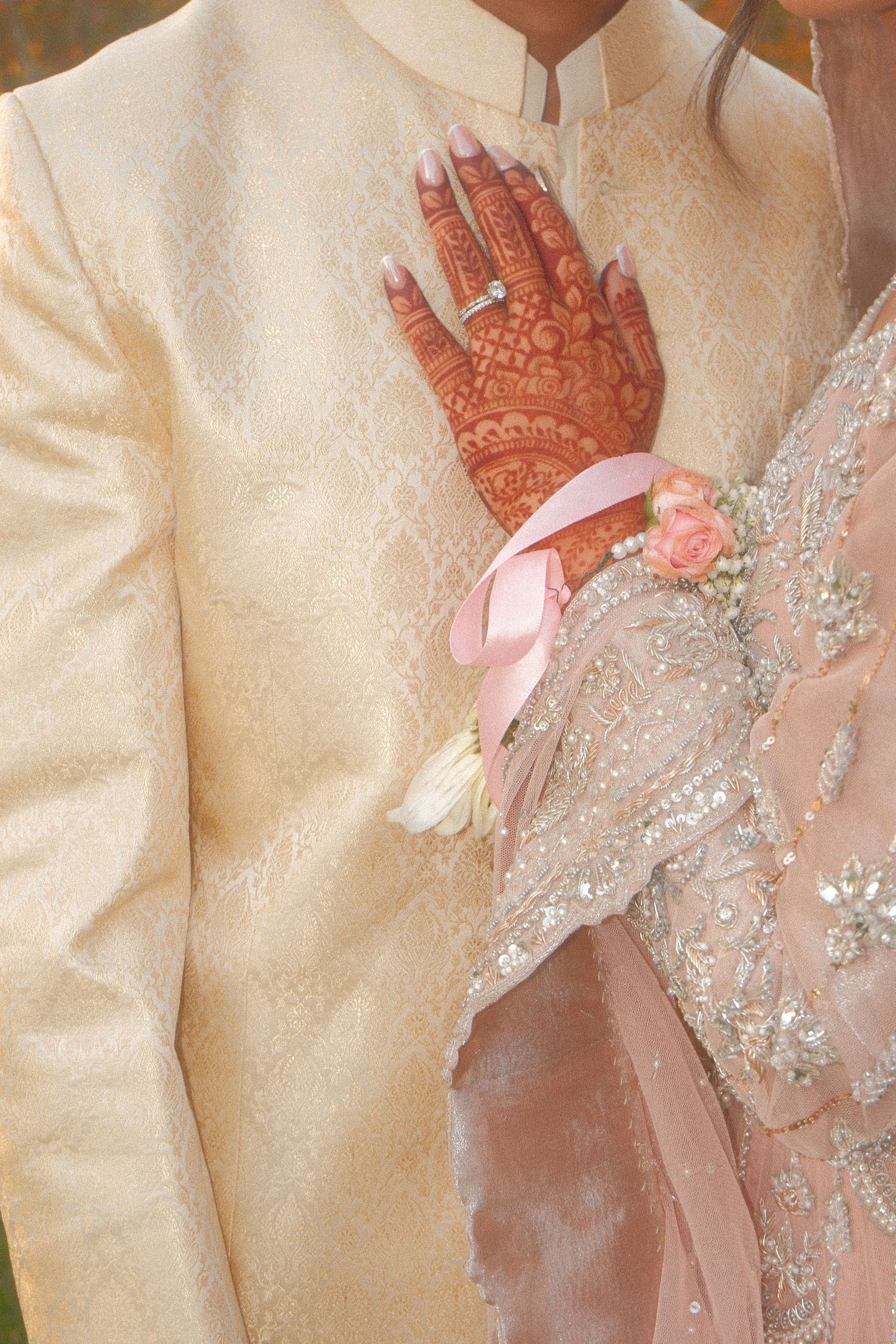 Close-up of South Asian bride with mehndi henna hands holding groom in cream sherwani, with pink floral wrist corsage.
