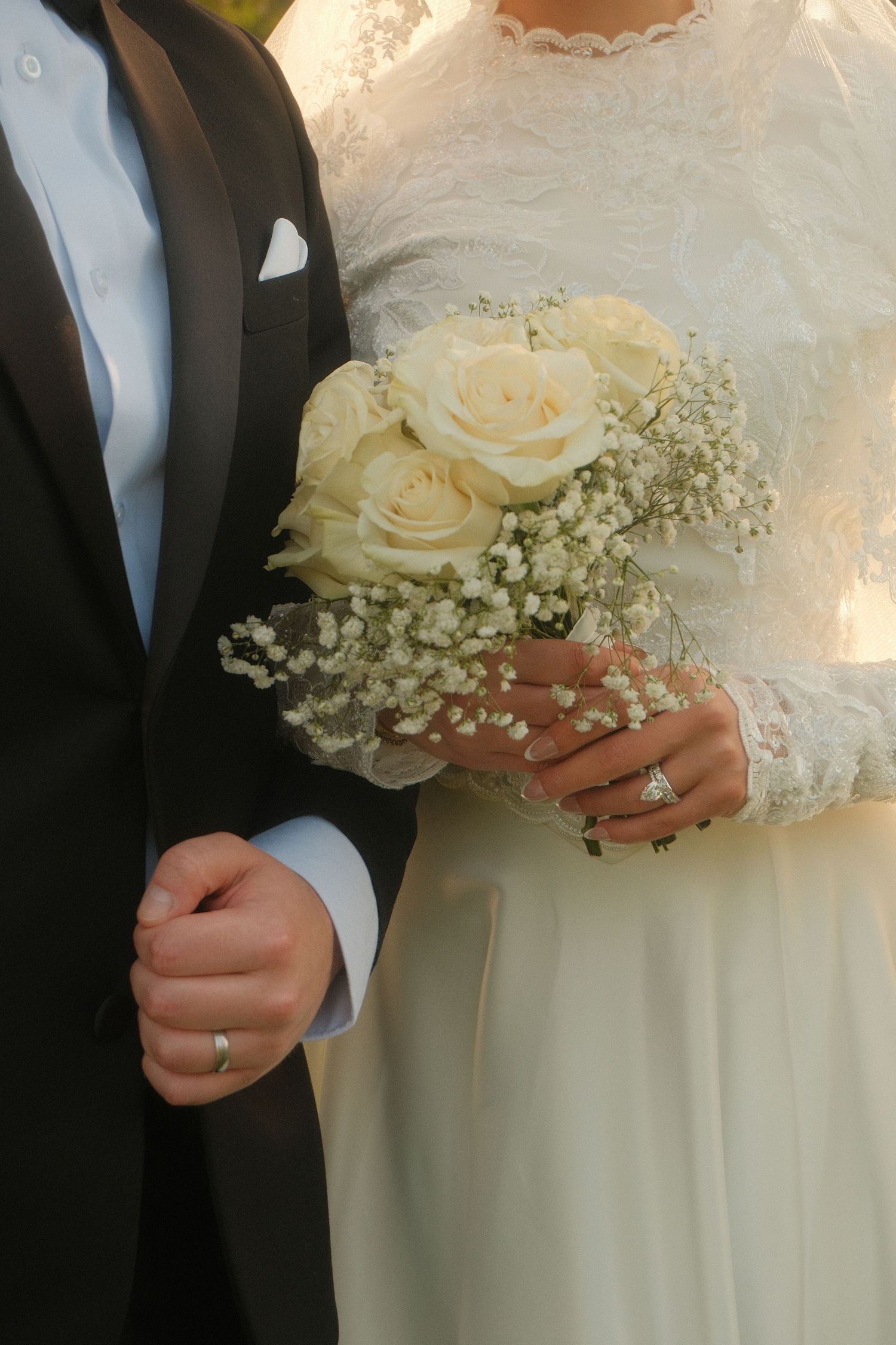 Bride and groom standing together, bride holding white rose and baby's breath bouquet, both wearing wedding rings.