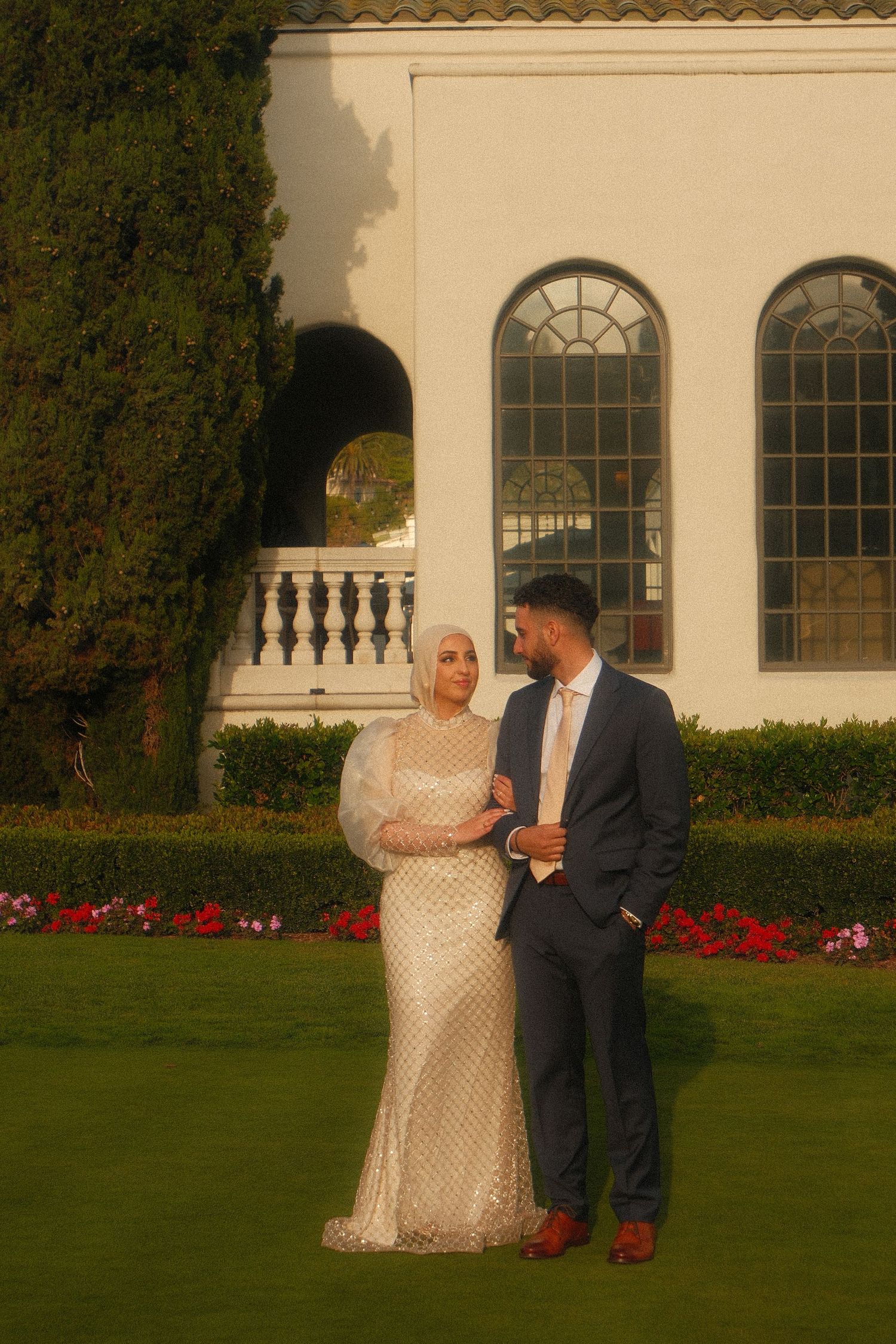 Elegant couple posing in formal attire on manicured lawn before a grand white villa with arched windows.