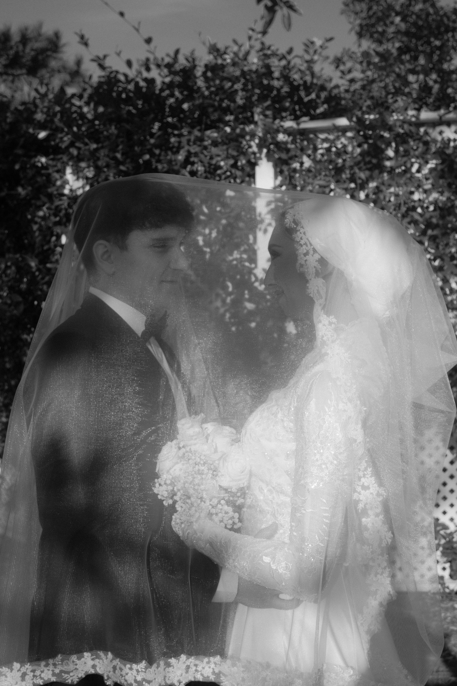 Black and white photo of bride and groom under veil at outdoor wedding ceremony.