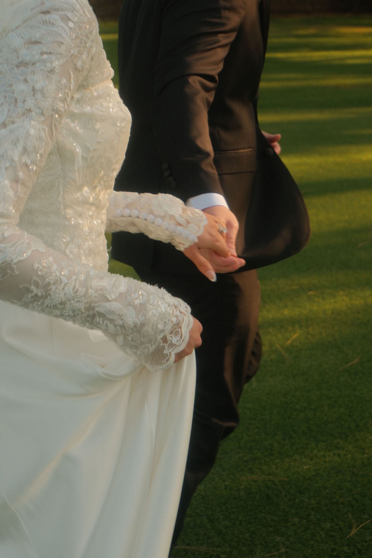 Bride in white lace gown and groom in dark suit holding hands on green grass at outdoor wedding.