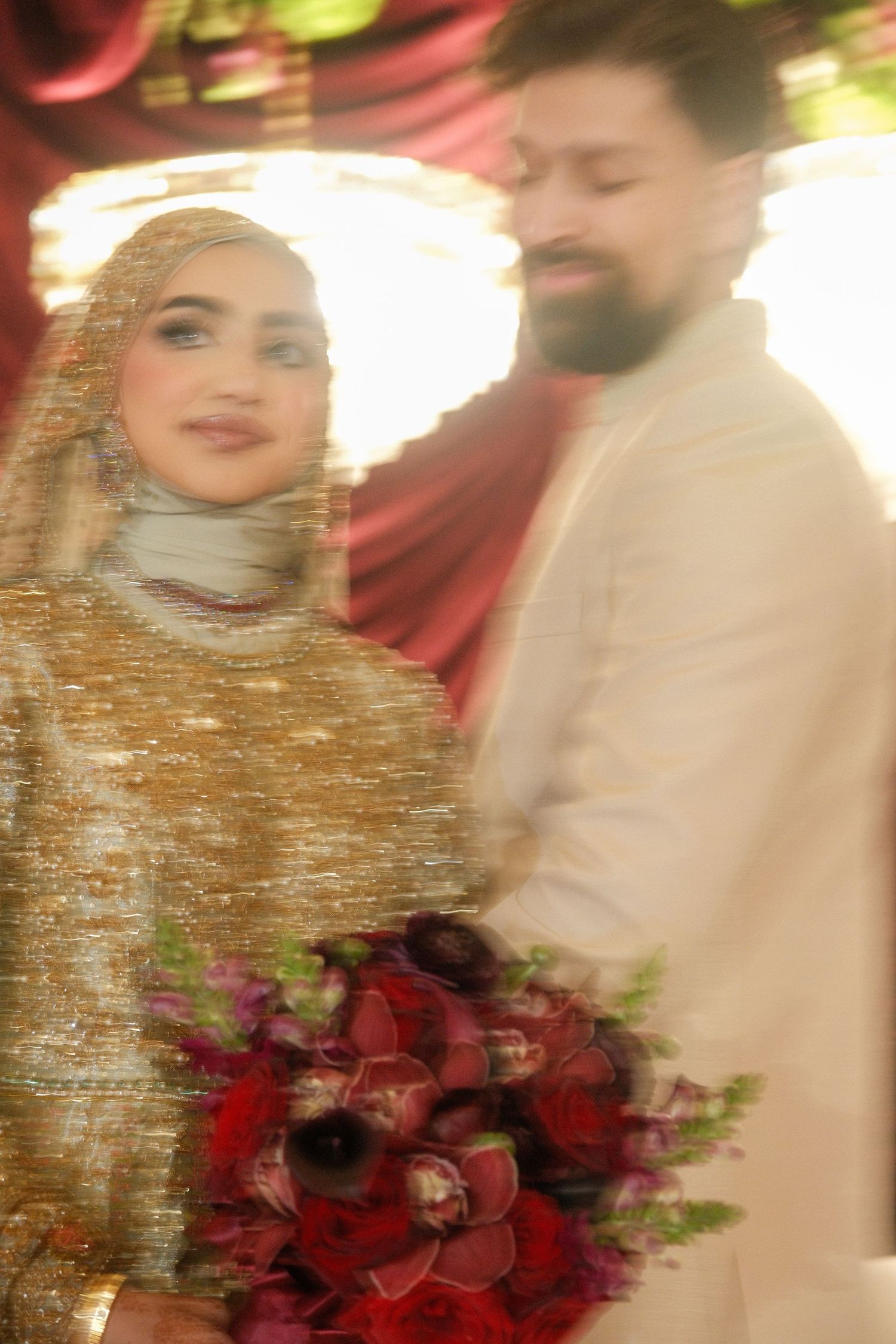Soft focus photo of Muslim bride in gold embroidered hijab holding red floral bouquet with groom beside her.