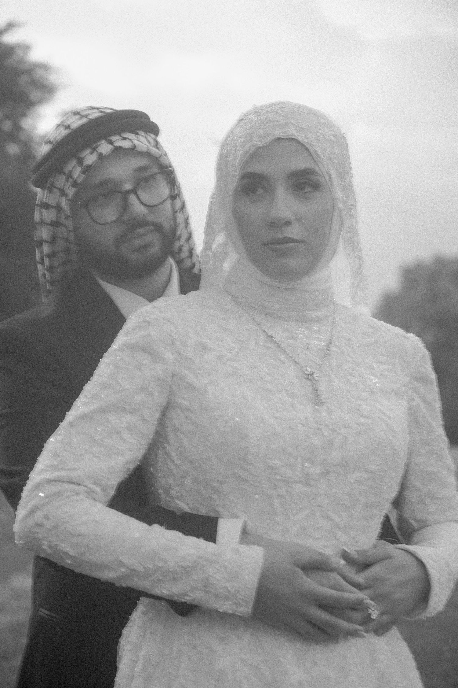 Black and white portrait of a Muslim bride in lace hijab gown with groom in keffiyeh embracing her.