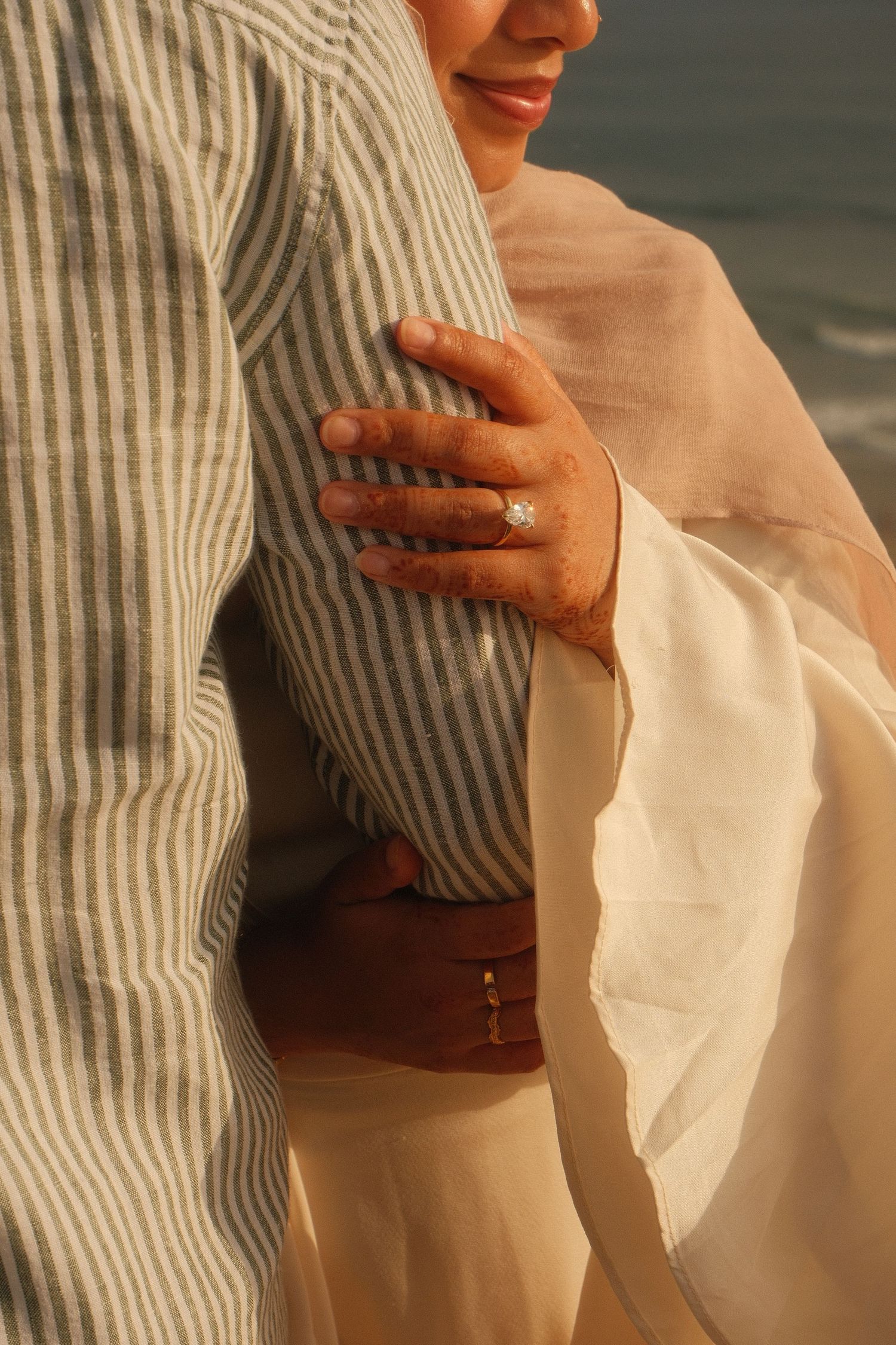Close-up of womans hand with diamond ring embracing man in striped shirt at golden hour beach setting.