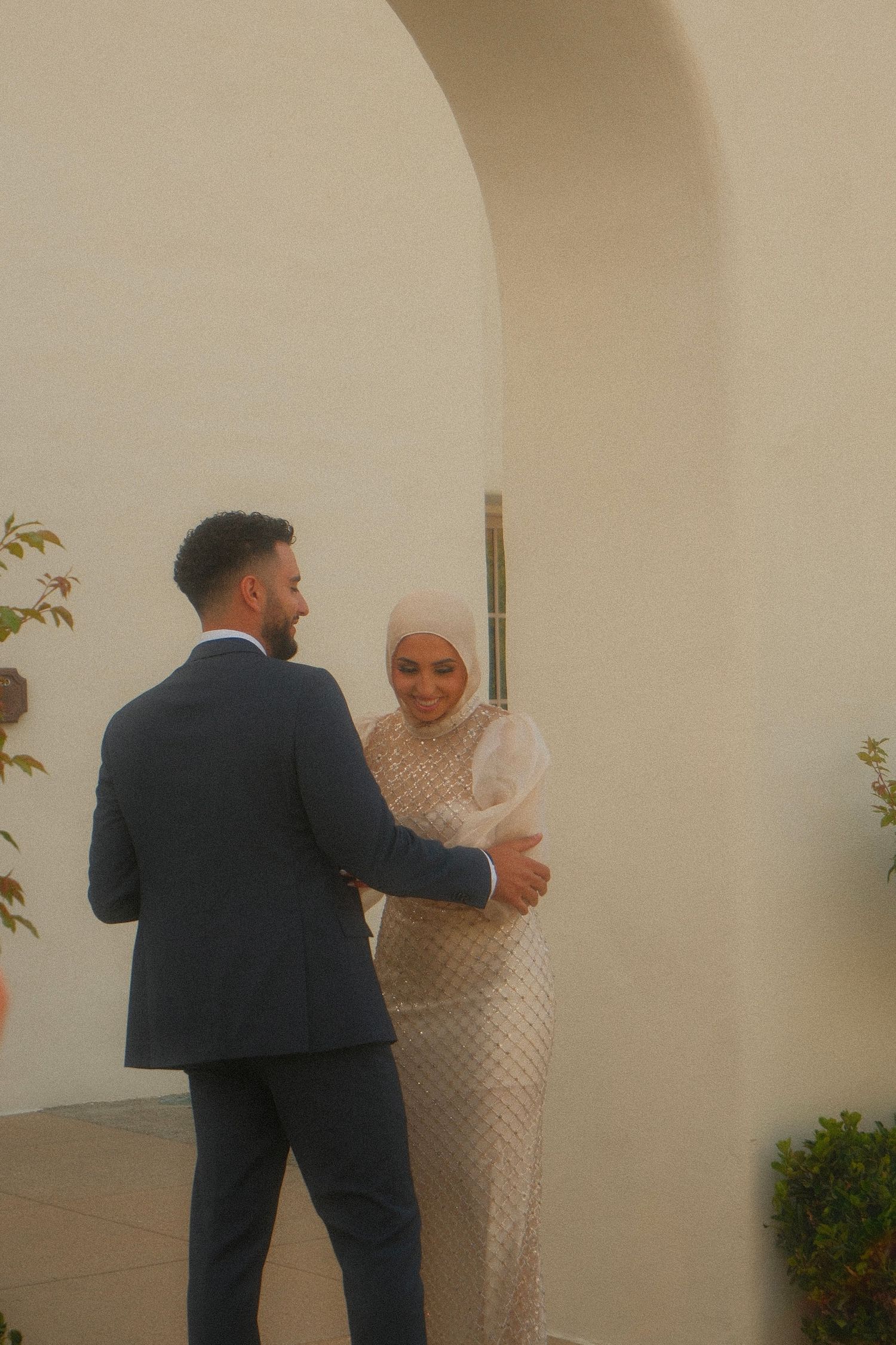 Muslim bride in embellished white dress and hijab sharing first look moment with groom in navy suit.