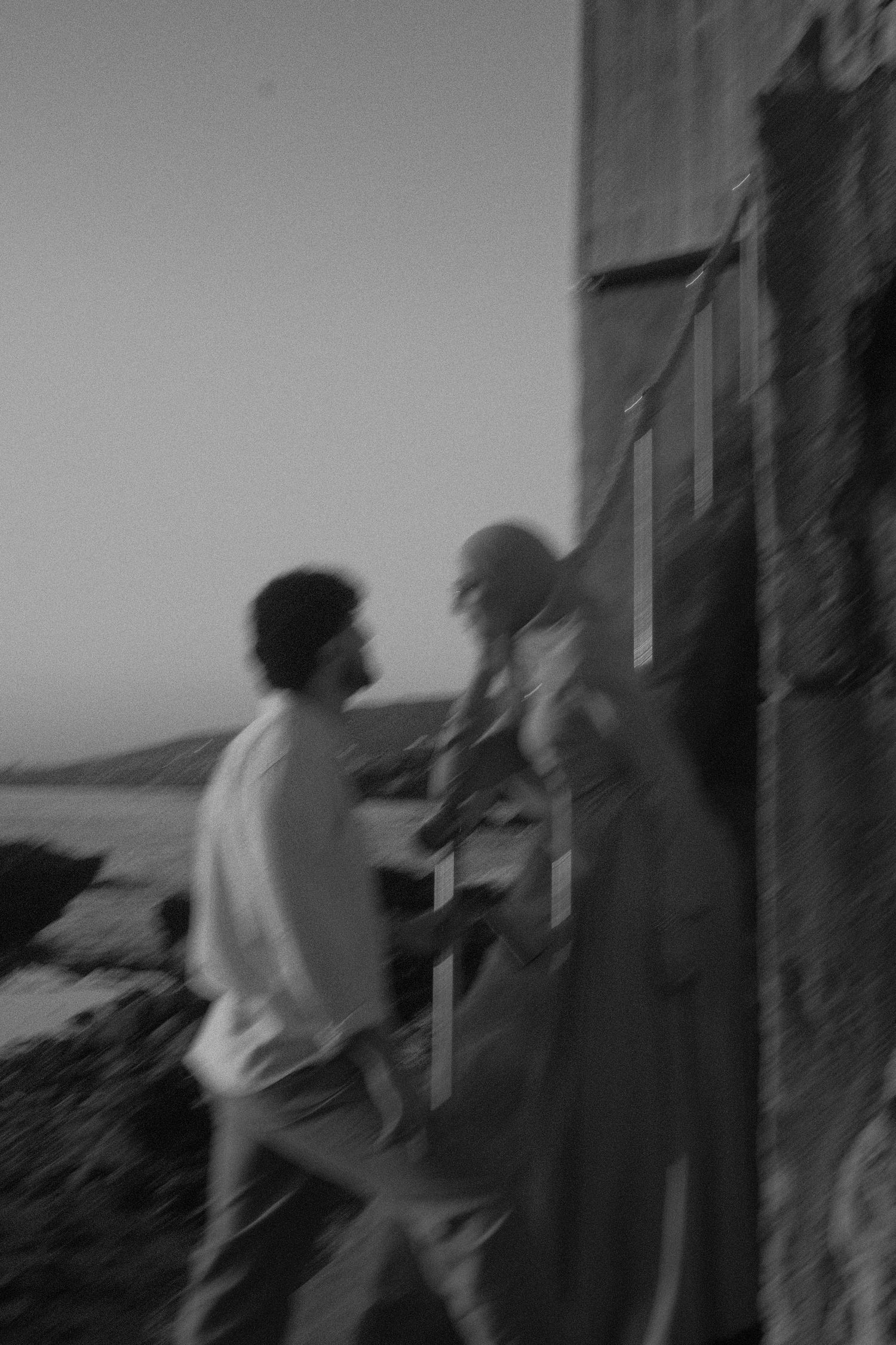 Black and white blurred motion photo of couple near stone wall at coastal sunset.