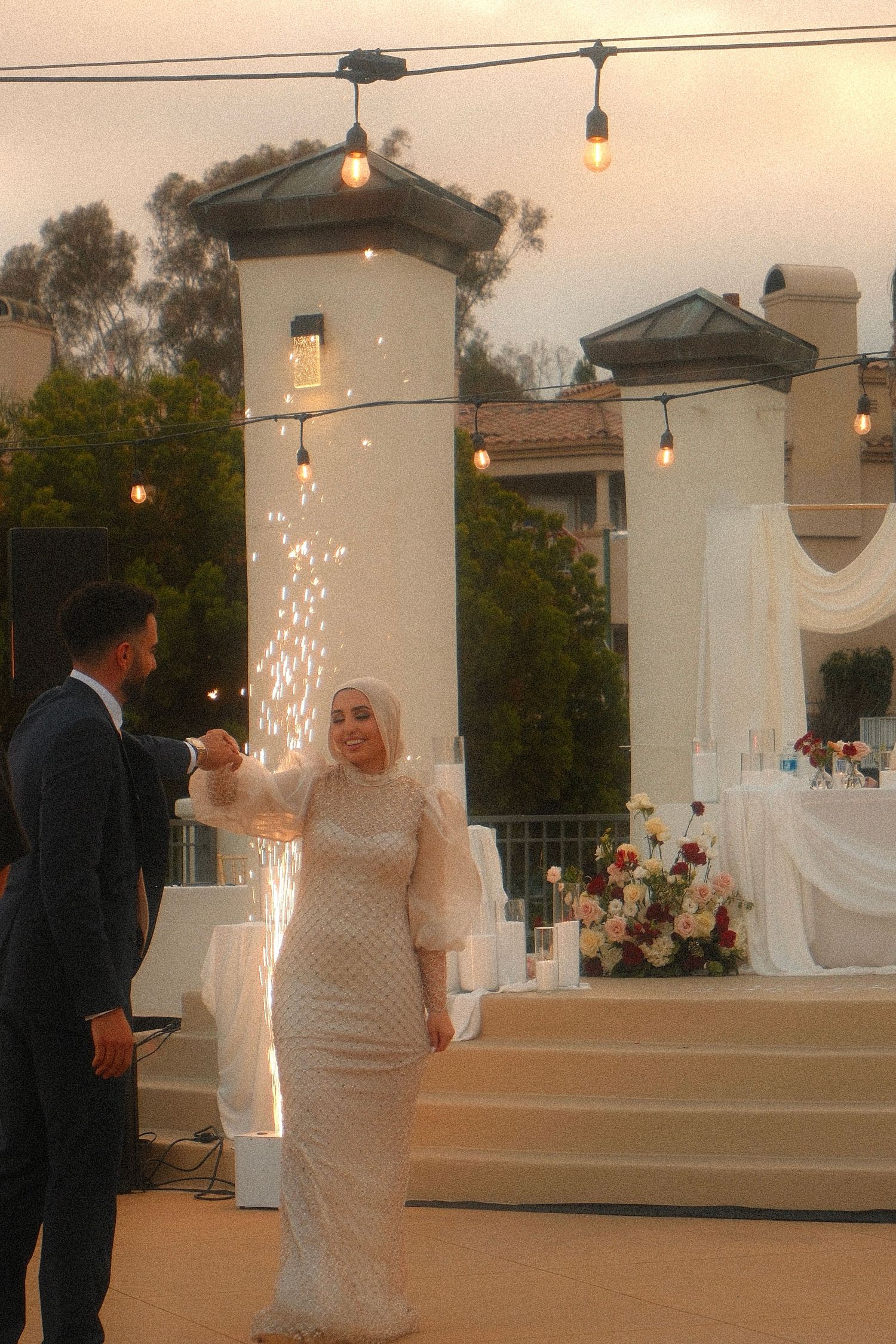Bride in white hijab and beaded gown holding sparkler at outdoor wedding reception with string lights.