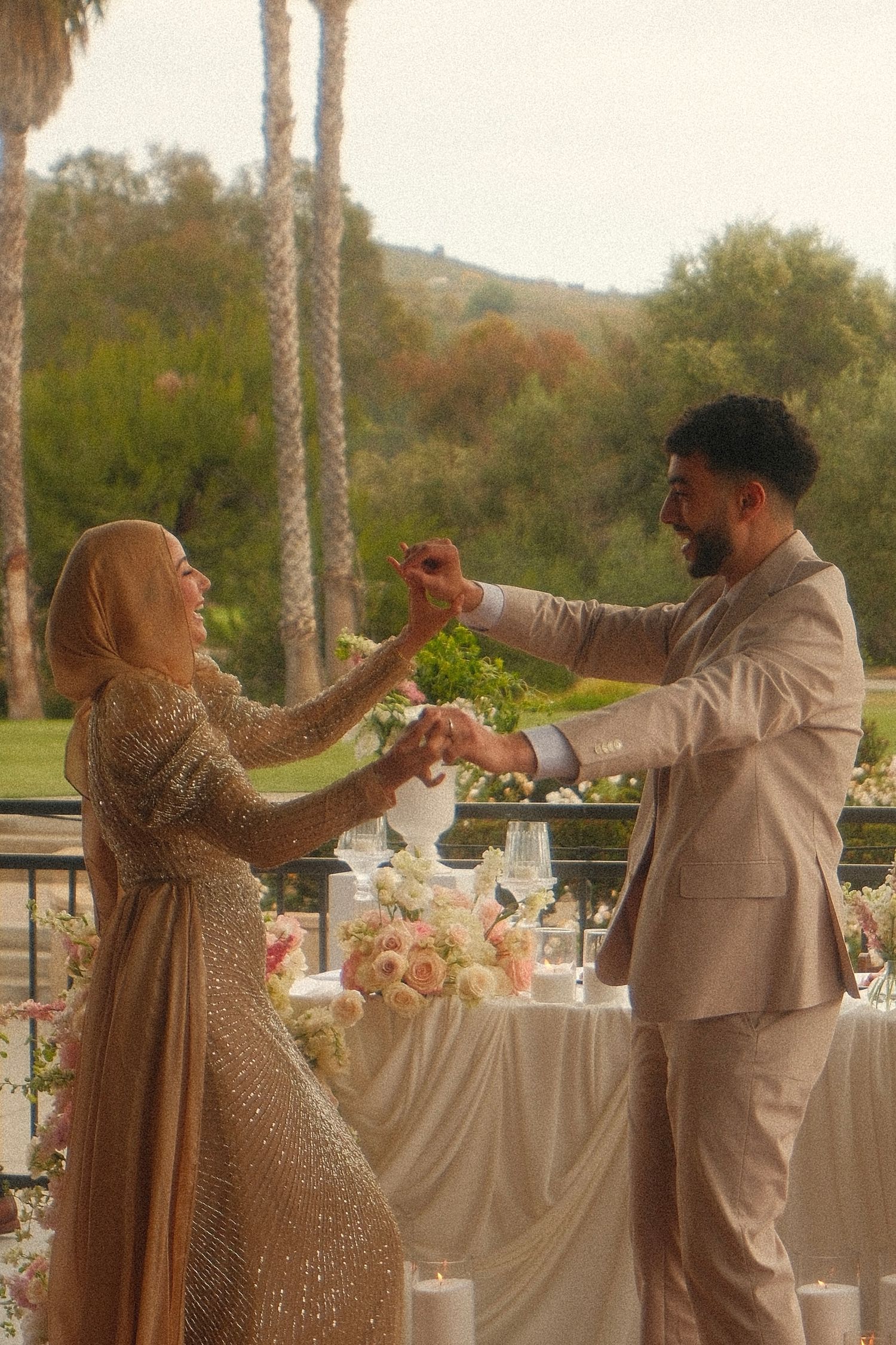 Muslim couple dancing outdoors at garden wedding reception, bride in gold sequin dress and hijab.