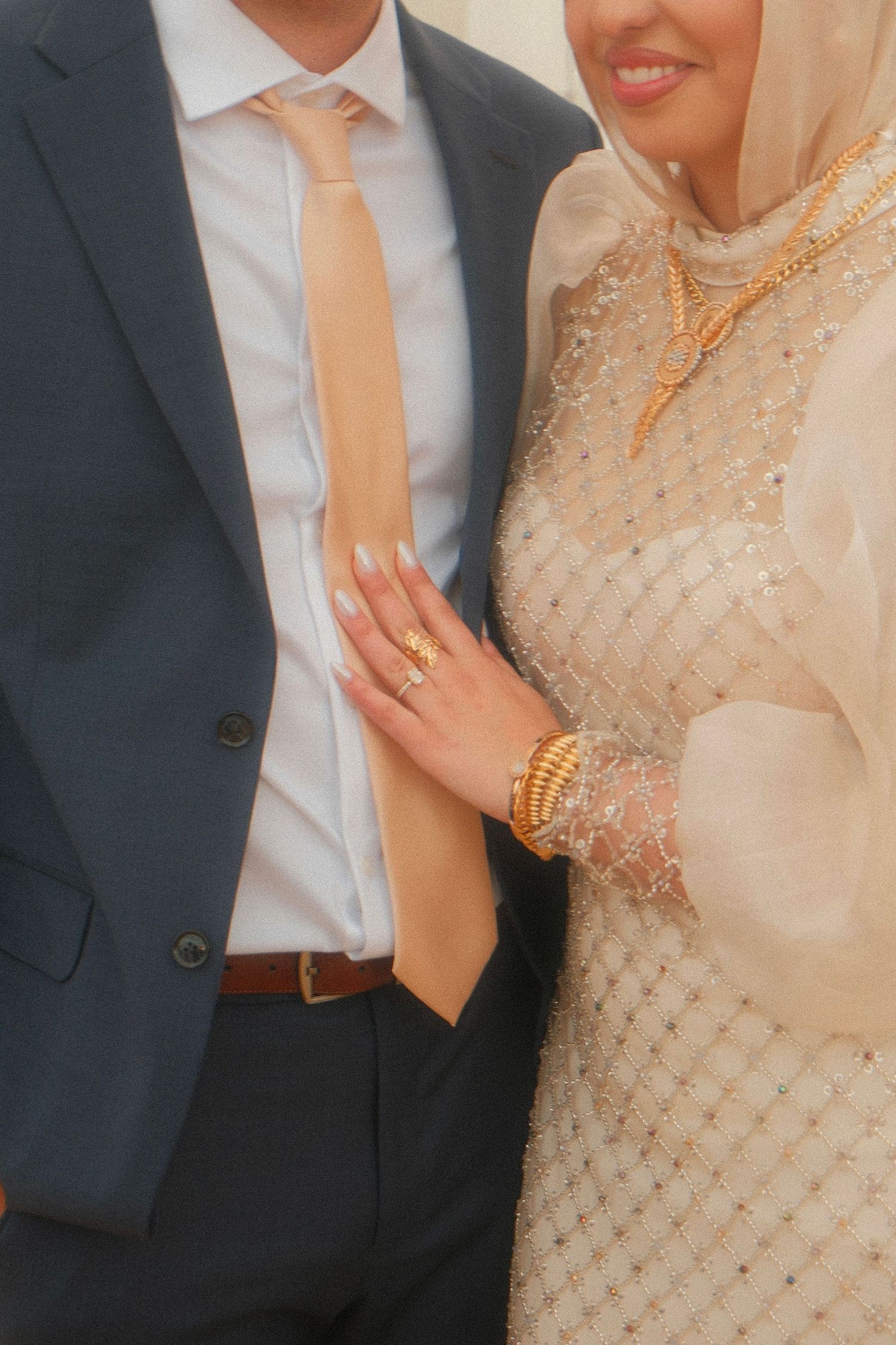 Close-up of bride in beige hijab adorned with gold jewelry resting hand on grooms navy suit jacket.