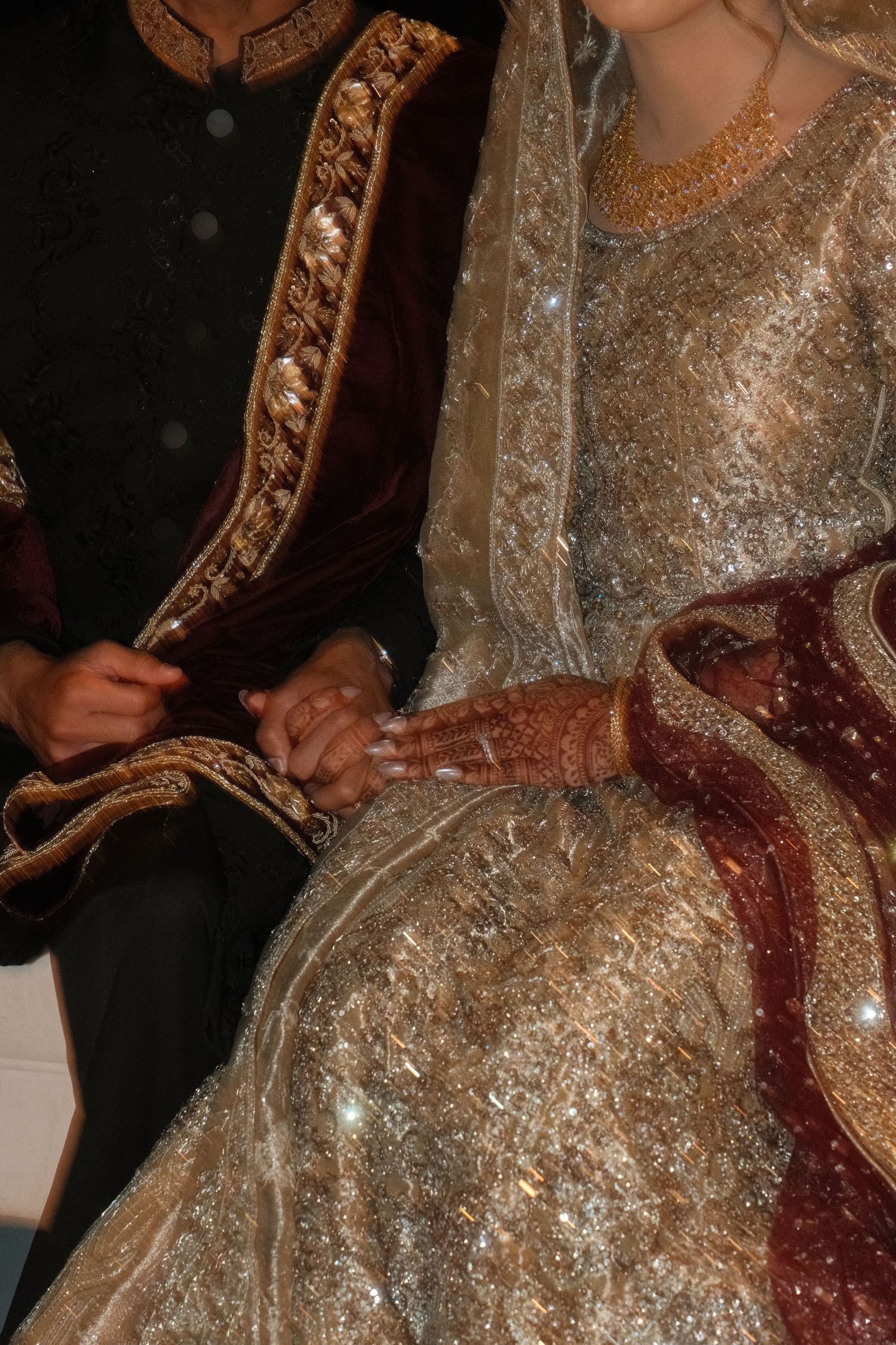 Close-up of South Asian wedding couple holding hands, bride in gold embroidered lehenga with mehndi, groom in dark sherwani.