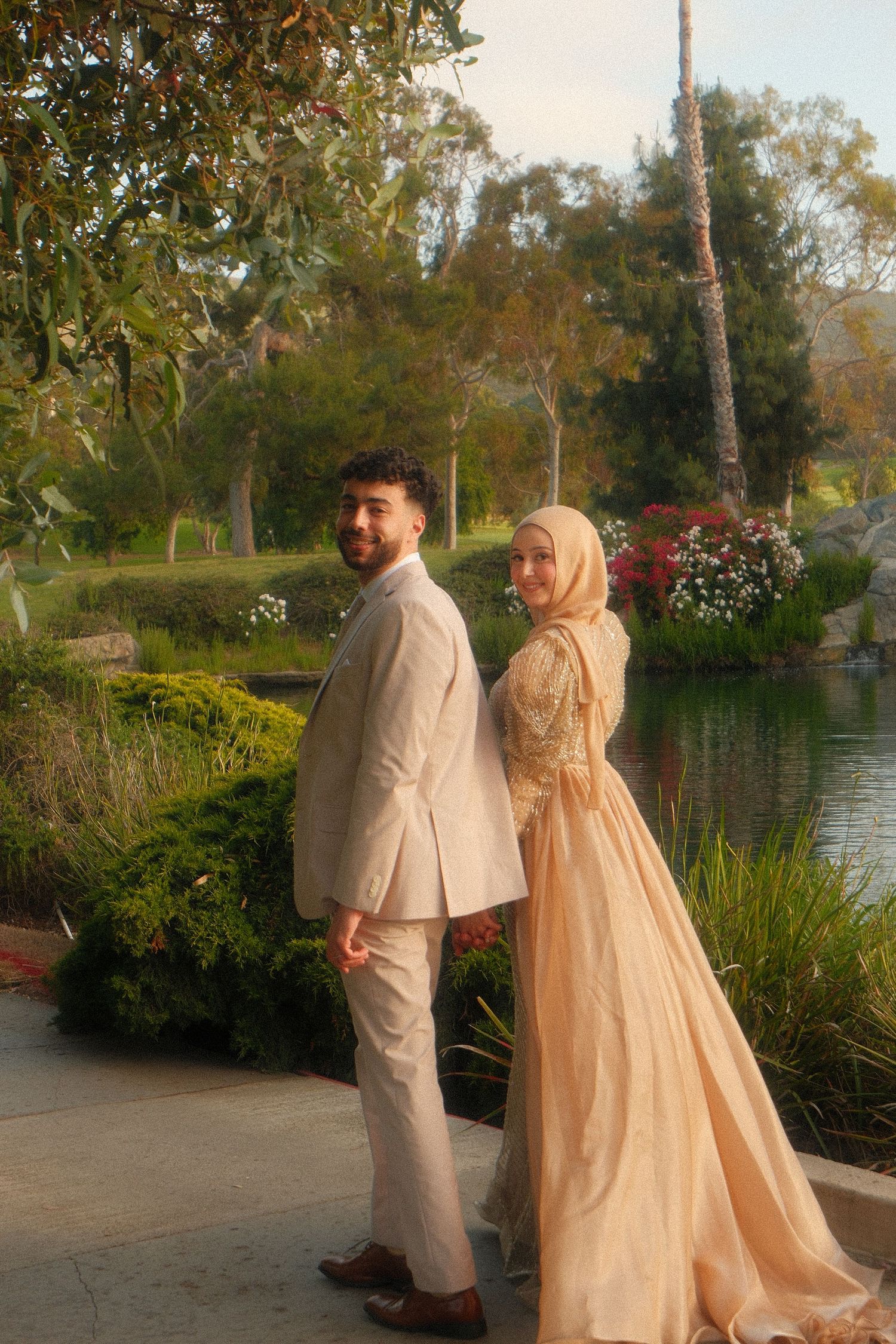 Muslim couple in elegant wedding attire posing by garden pond at golden hour.