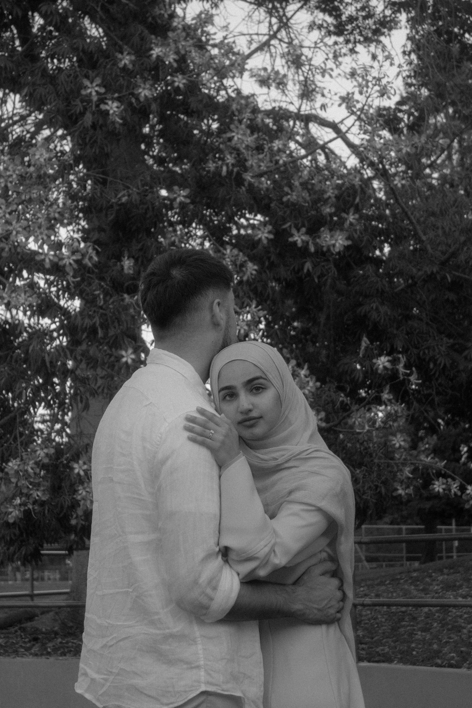Black and white photo of a couple embracing outdoors, woman wearing hijab smiling at camera near lush trees.
