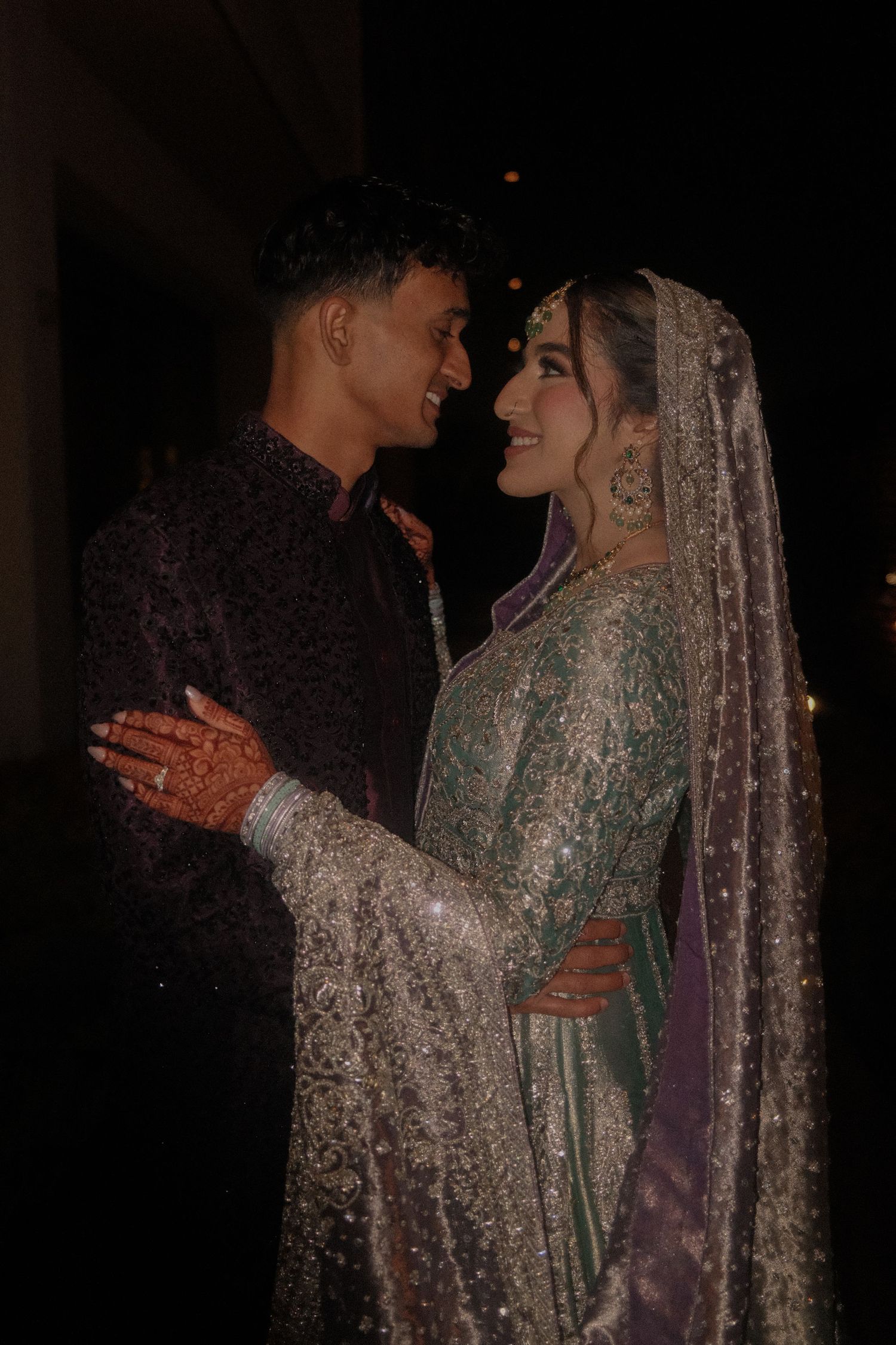 South Asian bride and groom sharing romantic moment at night, bride in teal silver lehenga with purple dupatta.