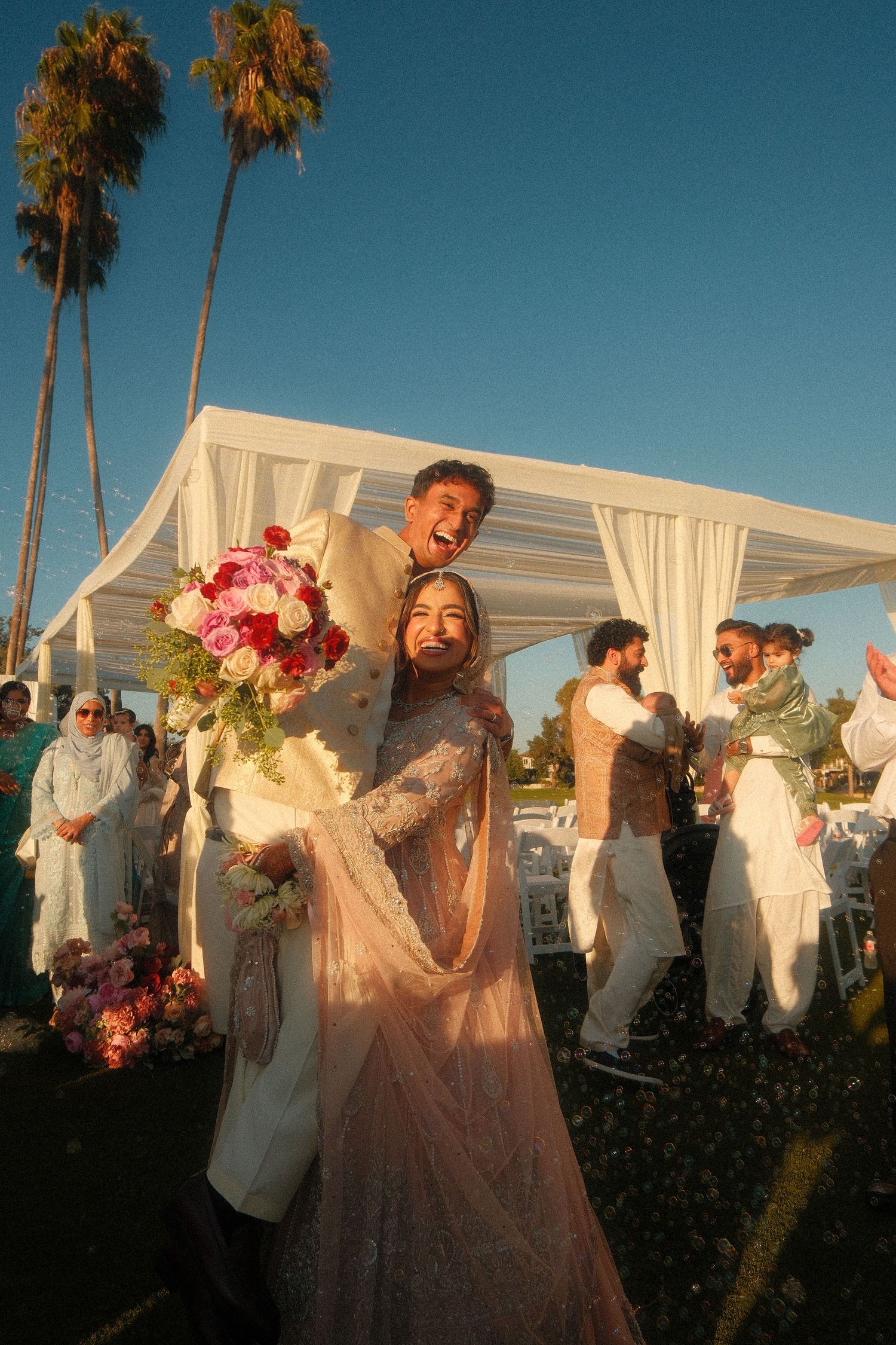 Joyful Muslim couple celebrating after their outdoor wedding ceremony under palm trees with pink bouquet.