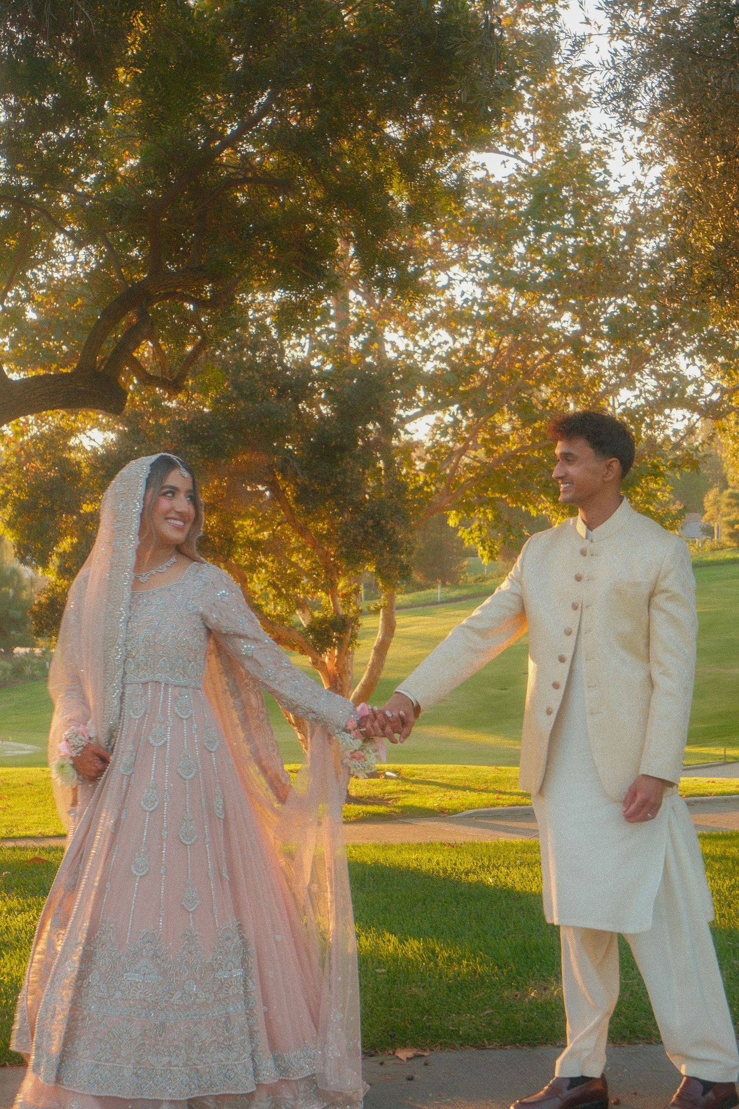 Pakistani wedding couple holding hands in golden sunset park, bride in pink lehenga and groom in cream sherwani.