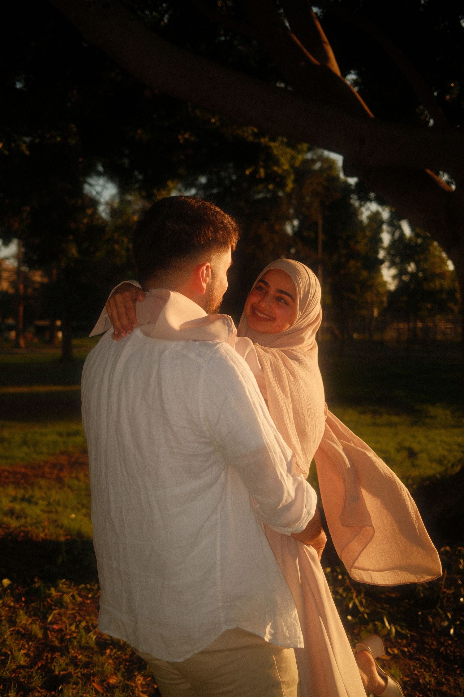 Couple sharing a warm embrace at sunset in a park, woman in beige hijab smiling over mans shoulder.